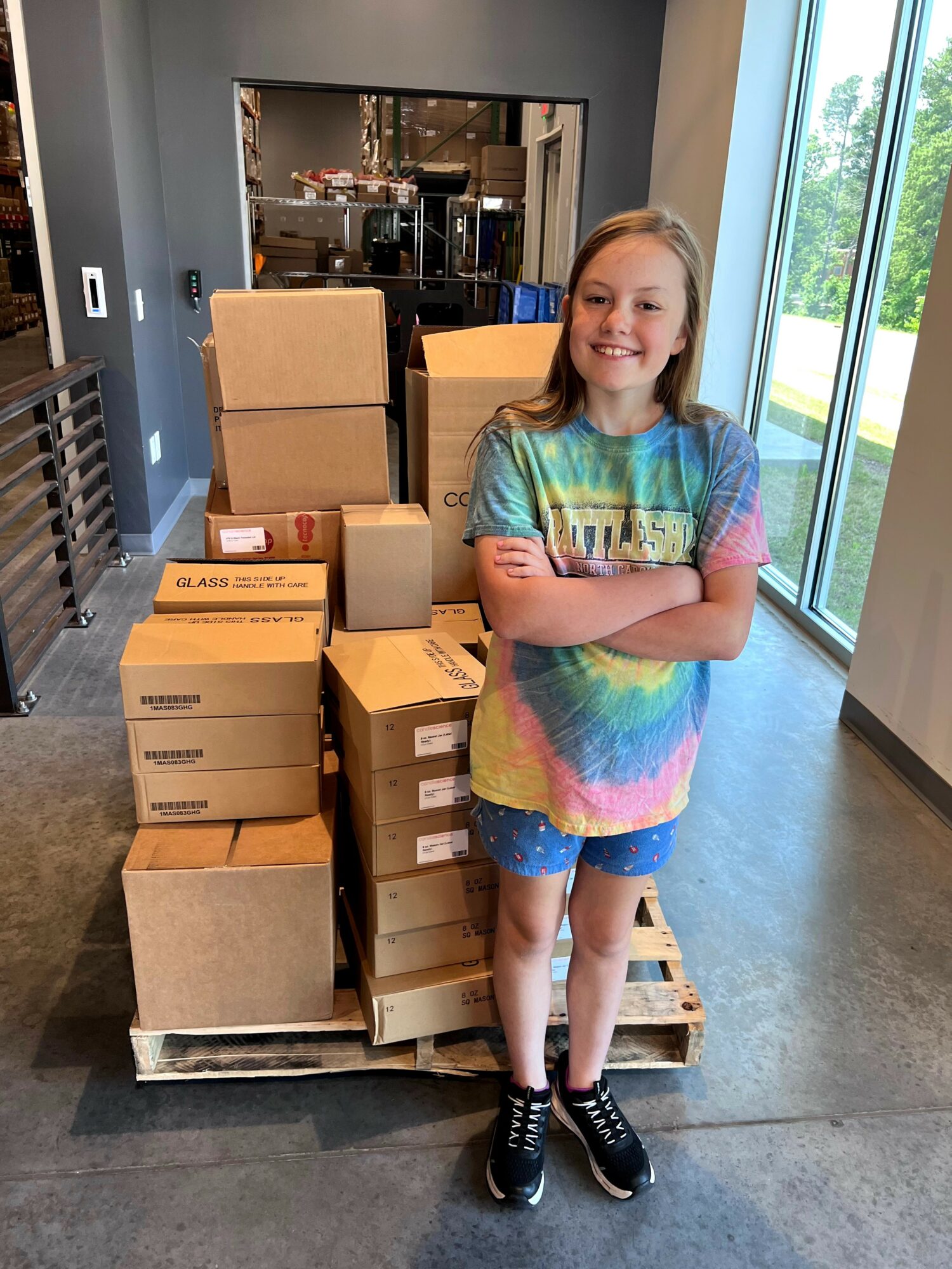 Smiling girl with crossed arms standing next to stacked boxes on a pallet in a room with large windows and a view outside.