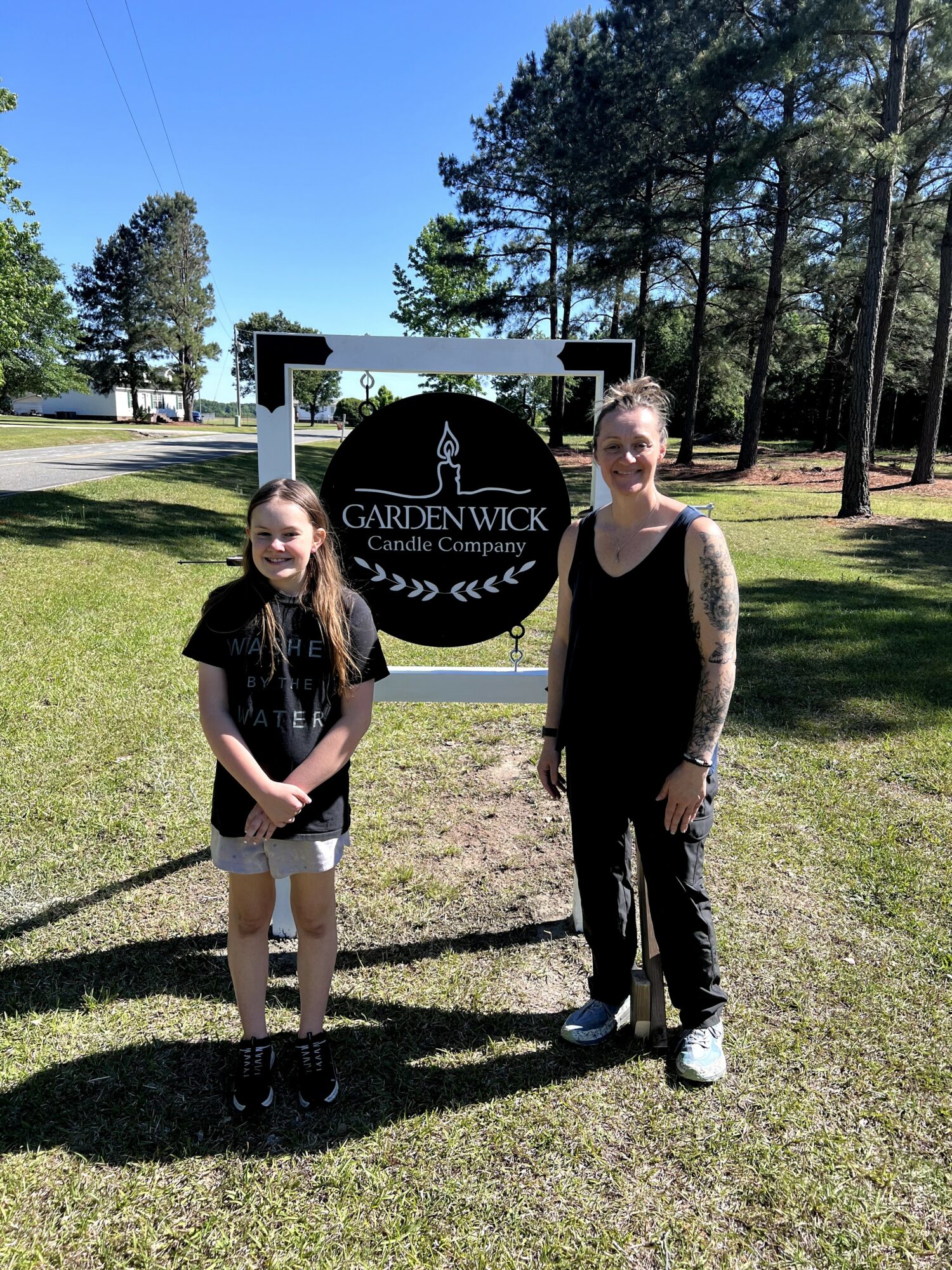 Two people standing outdoors in front of a sign for Gardenwick Candle Company, with trees and a blue sky in the background.