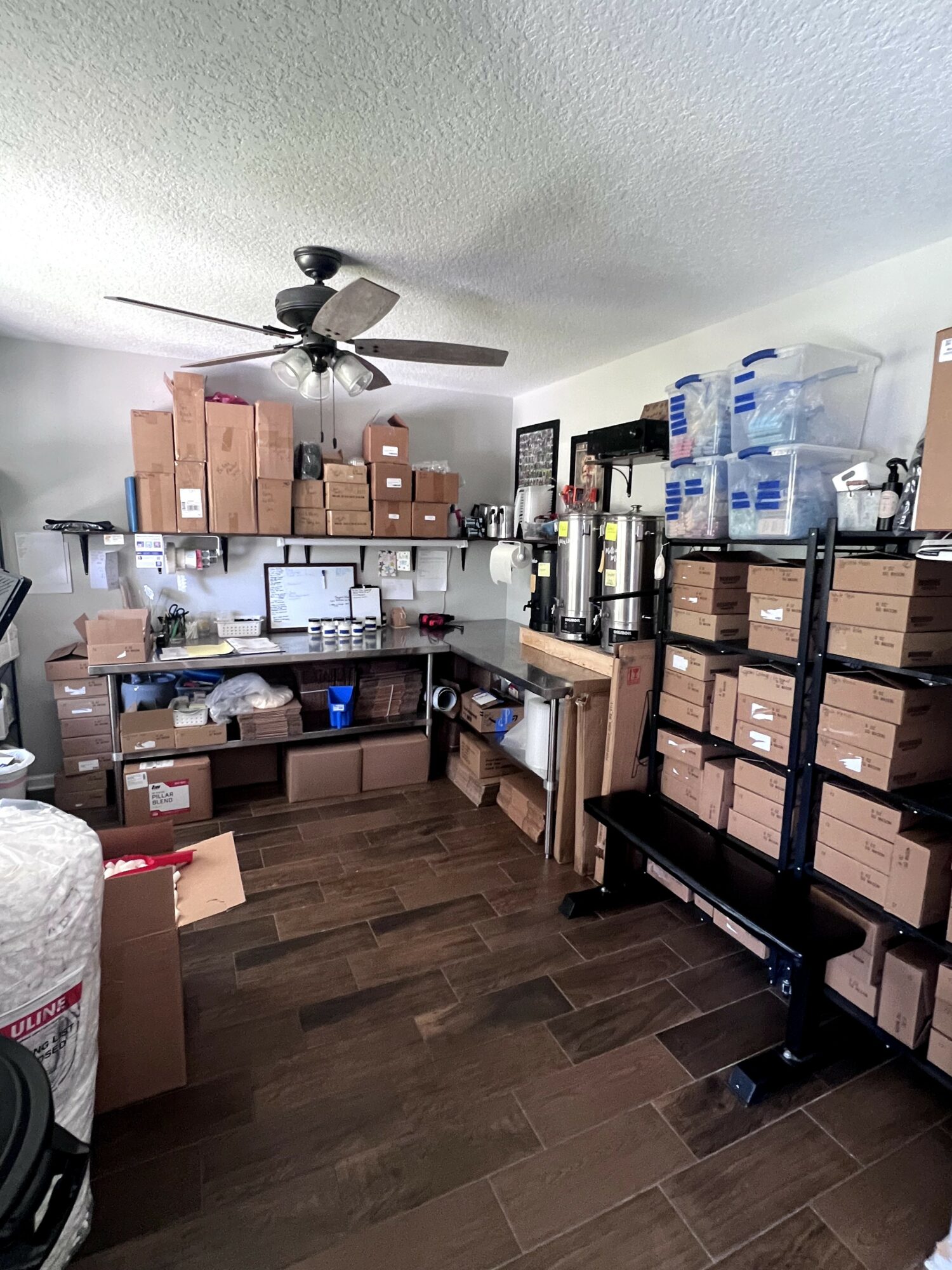 Room with shelves filled with boxes, supplies, and water bottles, ceiling fan, and wooden floor.