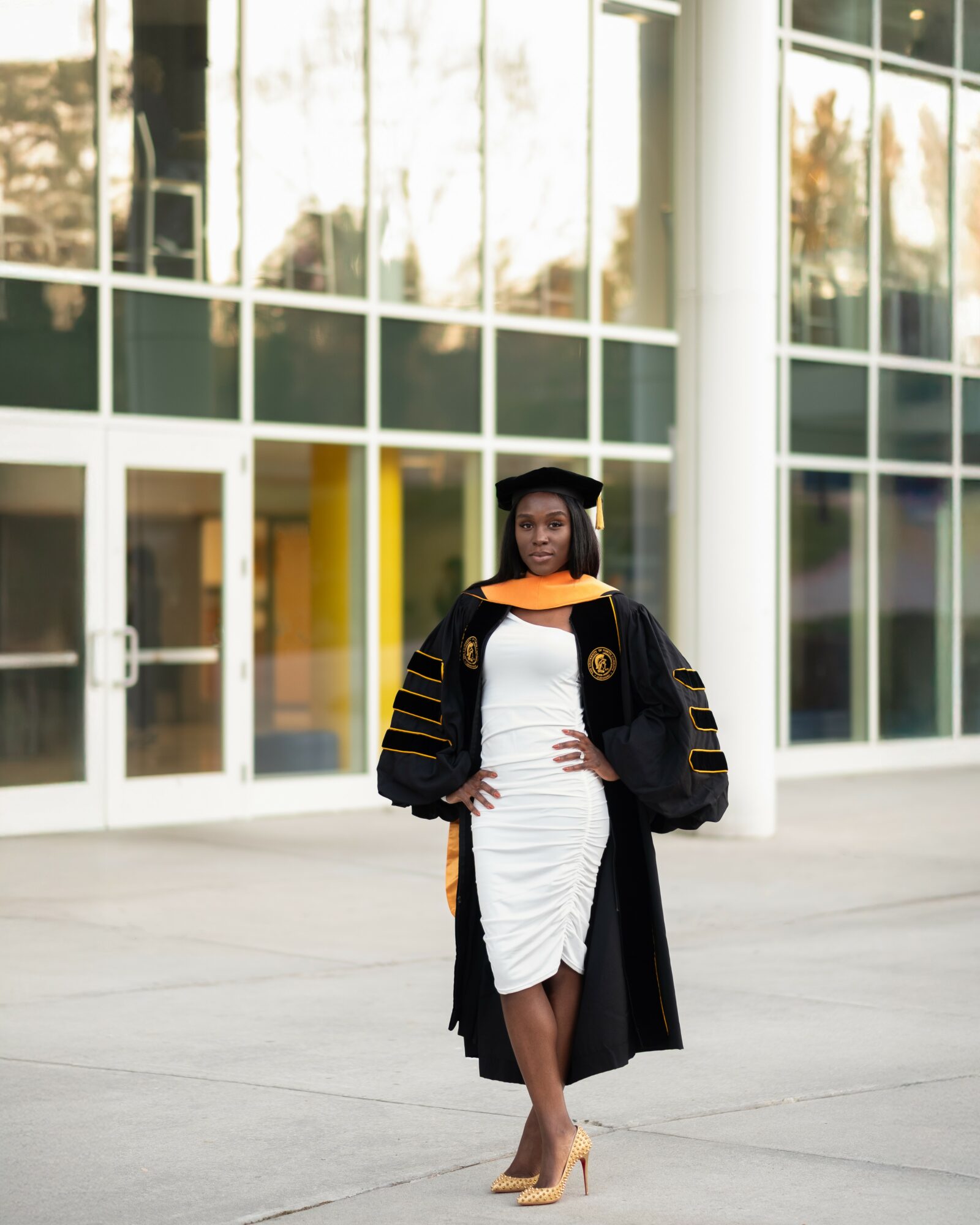 Woman in graduation gown and cap standing outside in front of glass building, wearing a white dress and heels.