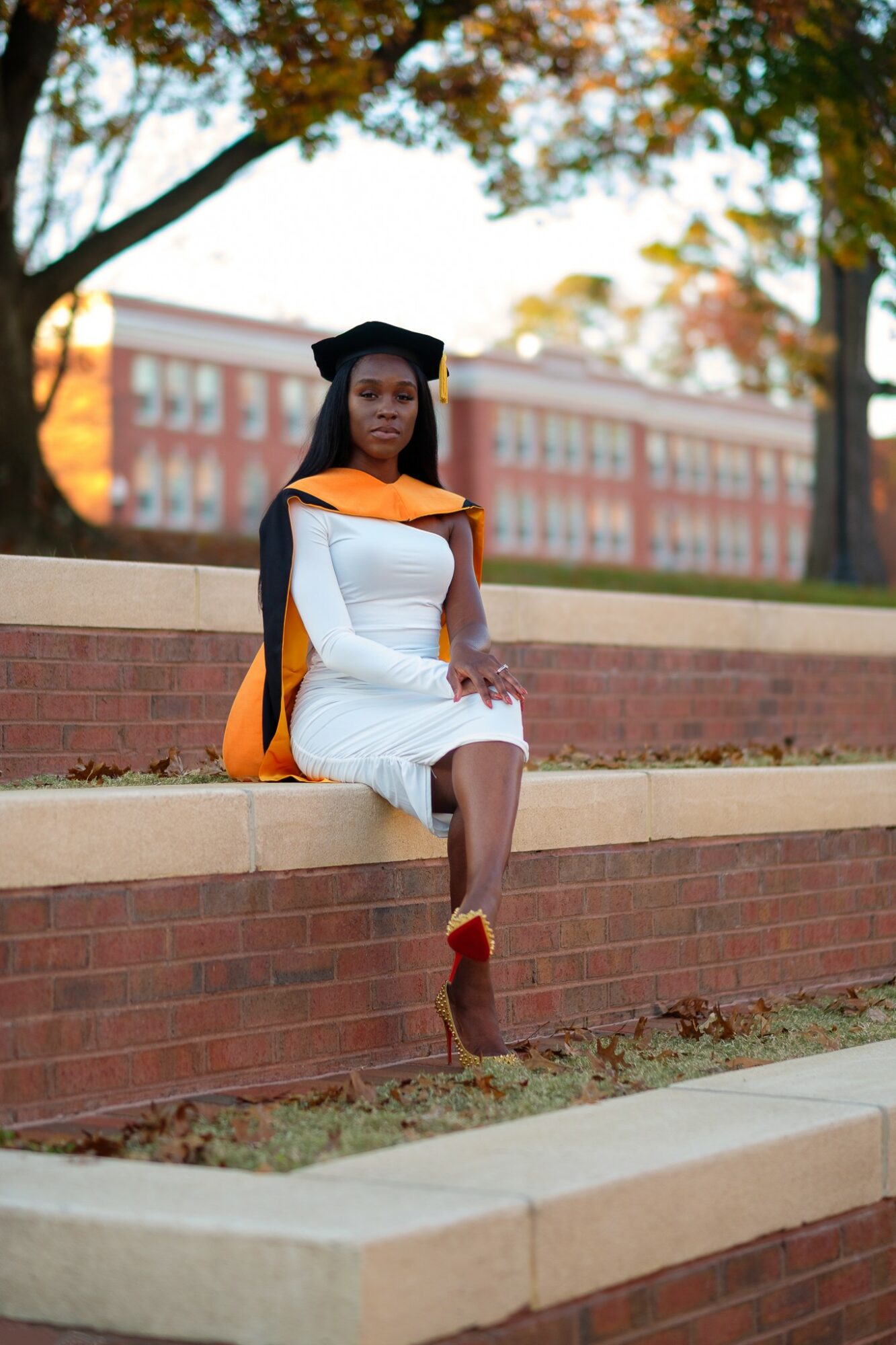 Woman in graduation cap and gown sitting on brick ledge outdoors, trees and building in background.