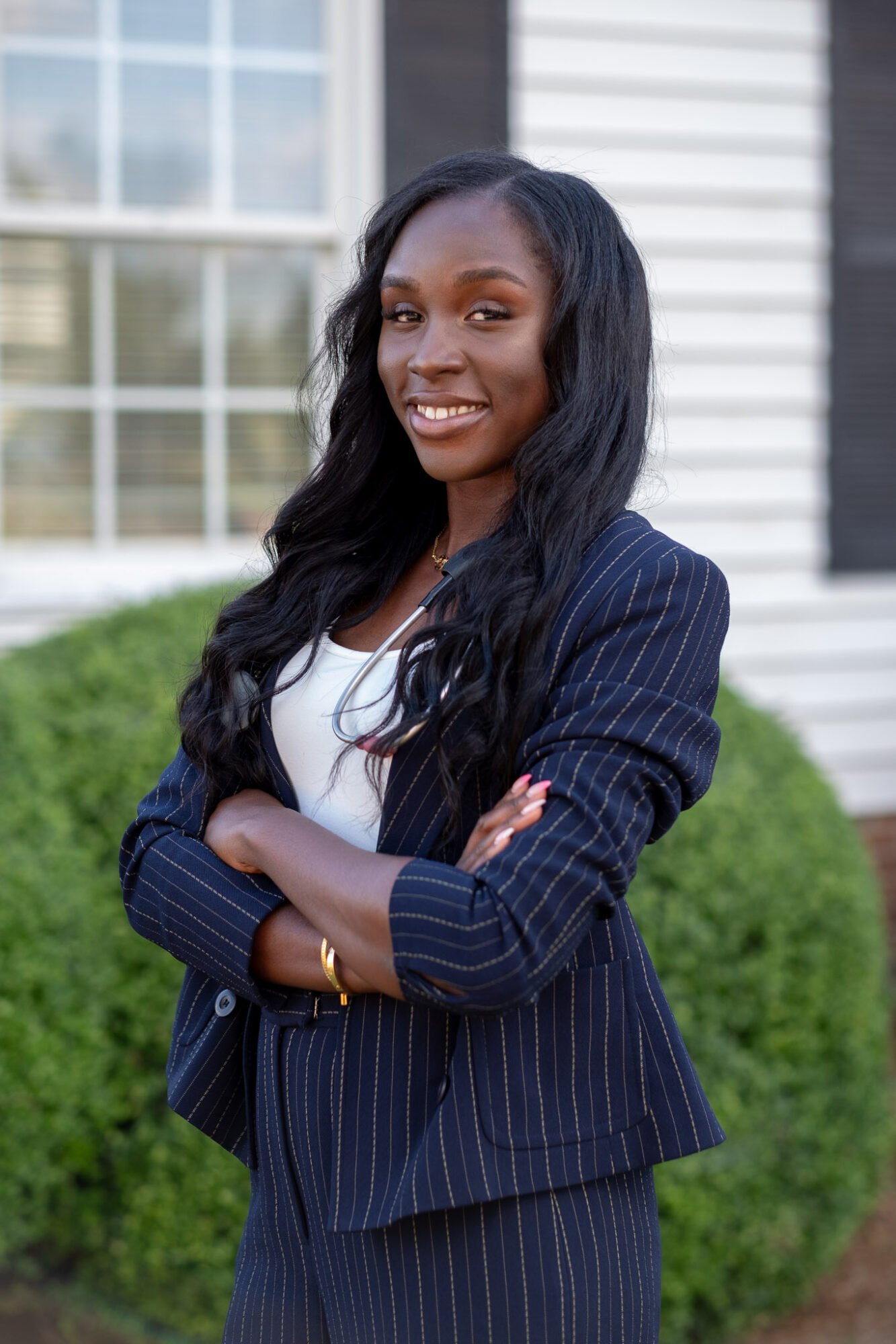 Smiling woman with long dark hair in a pinstripe suit outdoors, arms crossed, in front of a building and greenery.