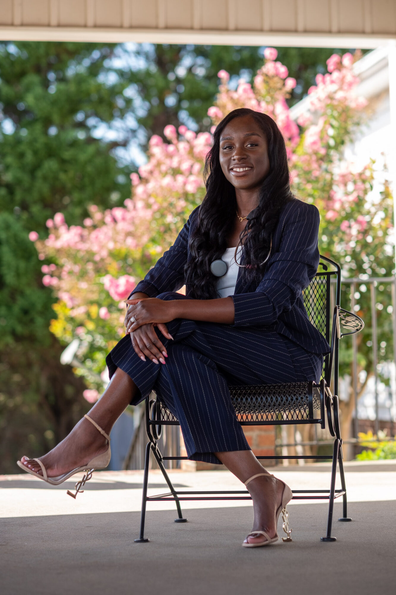 Woman sitting on a chair outdoors with pink flowering trees in background, smiling, wearing a dark pinstripe suit, heels, and sunglasses.