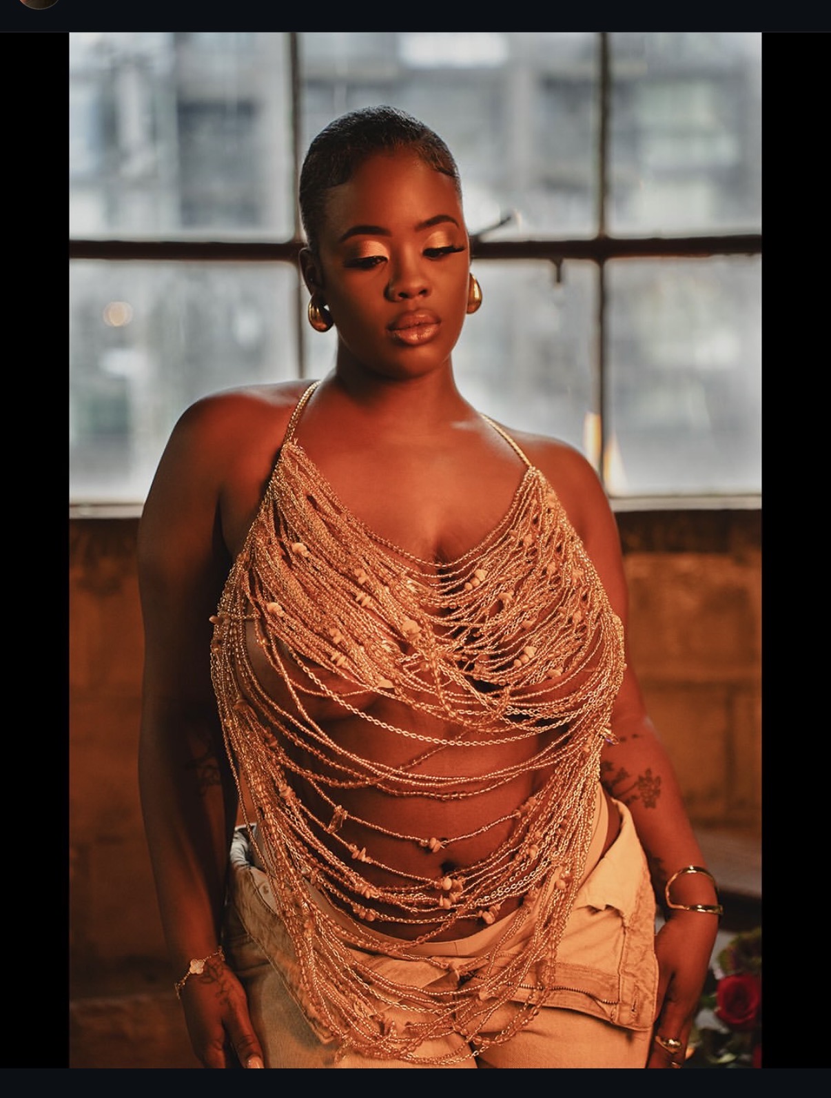 Young woman with braided hair wearing a beaded top and beige pants, standing indoors near a window.