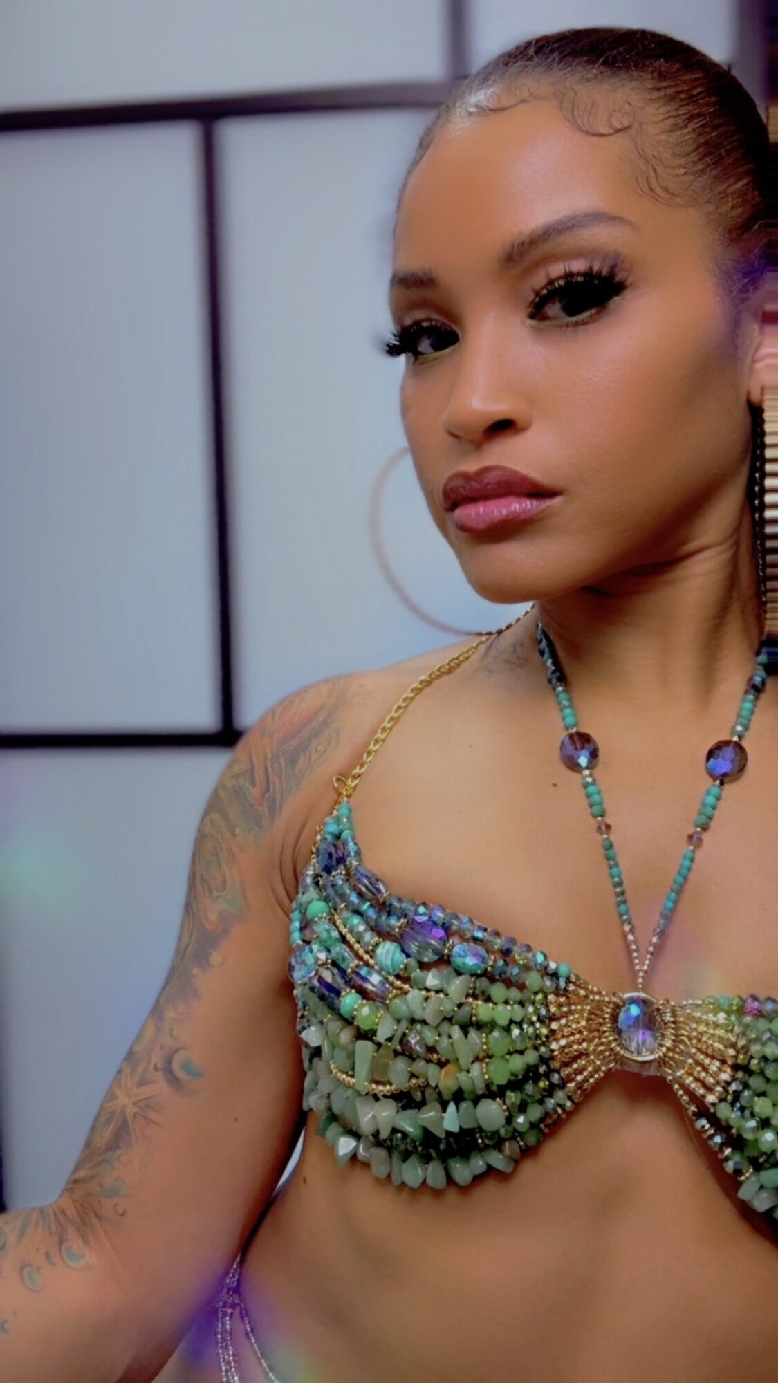 Woman with makeup and jewelry, wearing a beaded top, looking at the camera, background with geometric patterns.