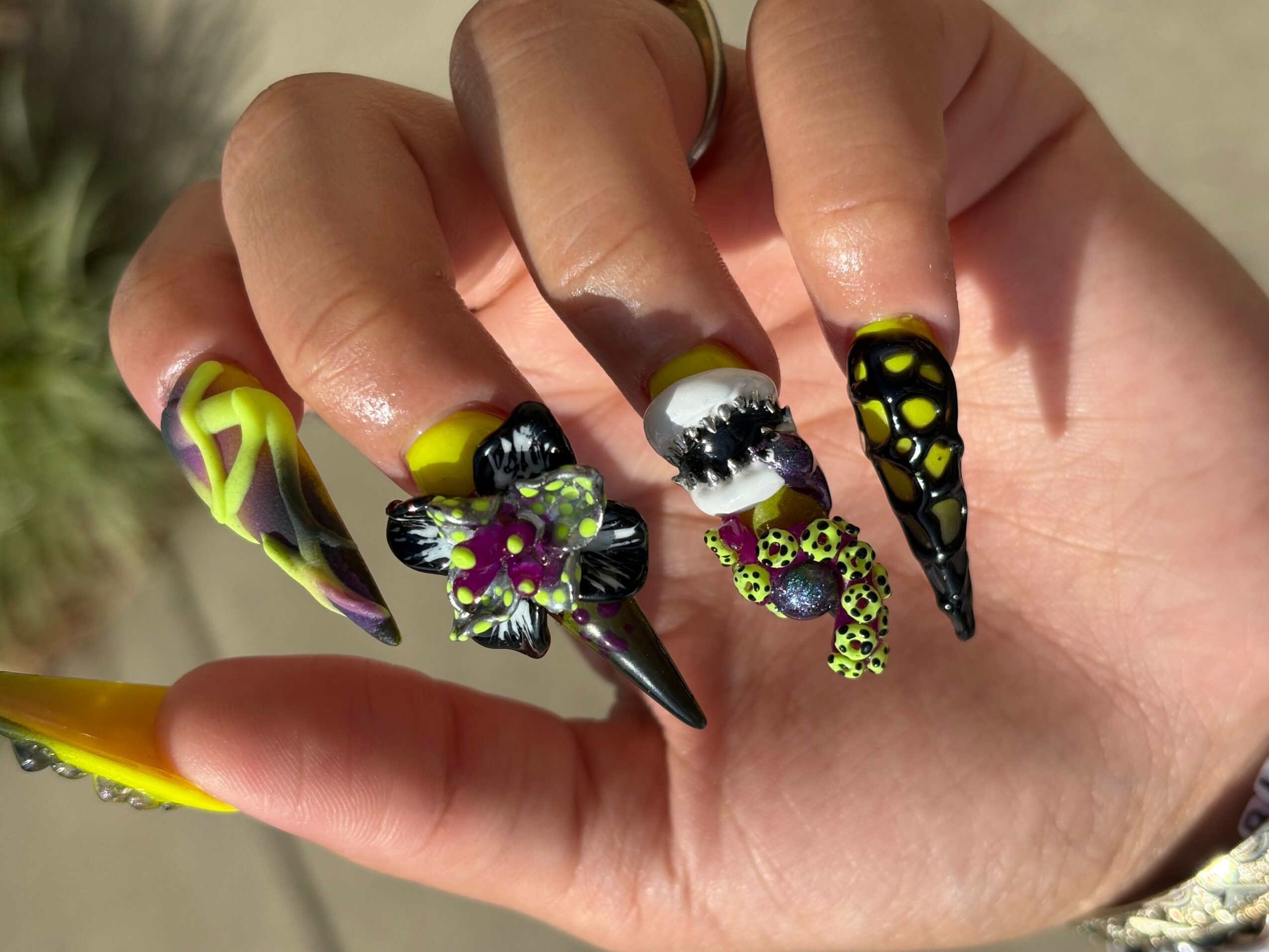Hand with long, decorated nails featuring black, yellow, and purple designs, including a butterfly and floral patterns.