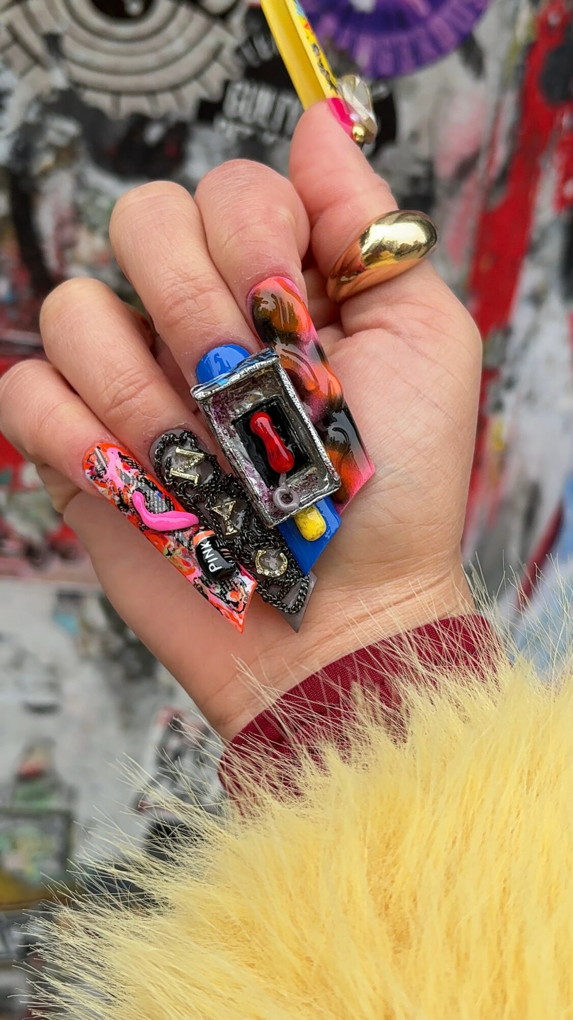 Hand holding a colorful toy with painted nails, wearing a yellow fuzzy sleeve and a large ring.