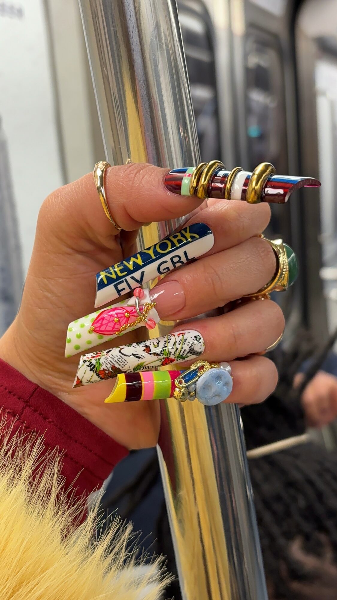 Hand holding a subway pole with colorful rings and a yellow bracelet reading 'New York Fly Girl,' with a blurred subway background.