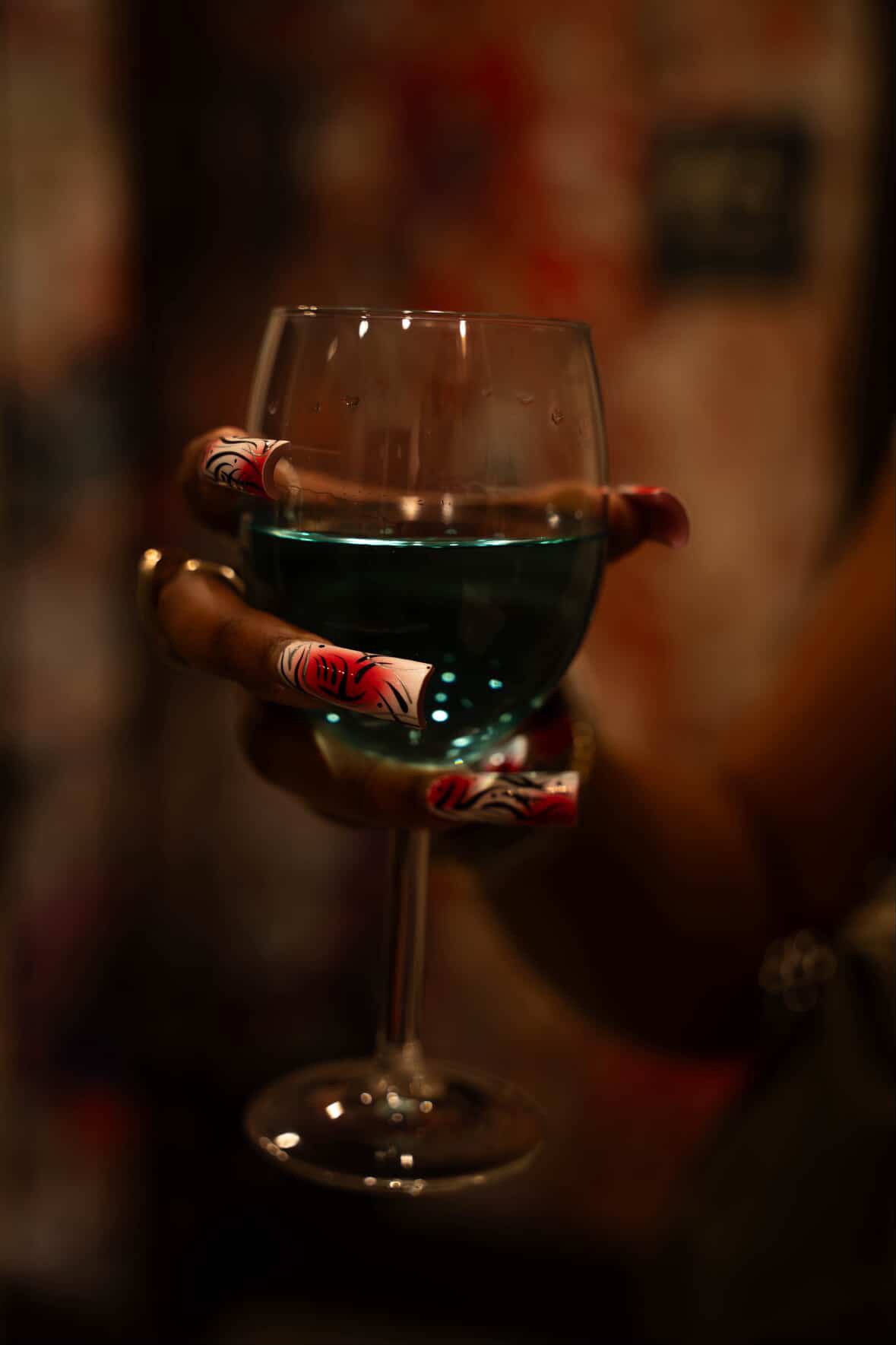 Person holding a glass of blue-green drink with decorative rolled paper around the stem, in a dimly lit setting.