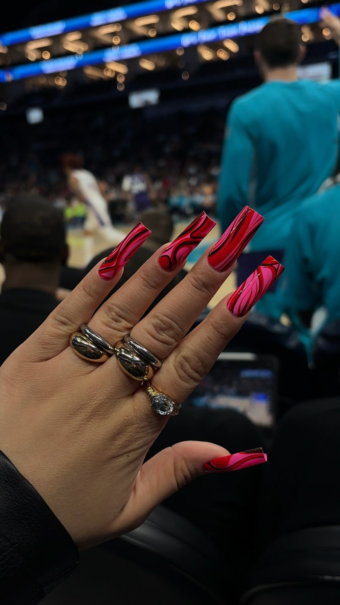 Hand with long pink nails and rings, in a crowded indoor event with people and a person in a blue jacket in the background.