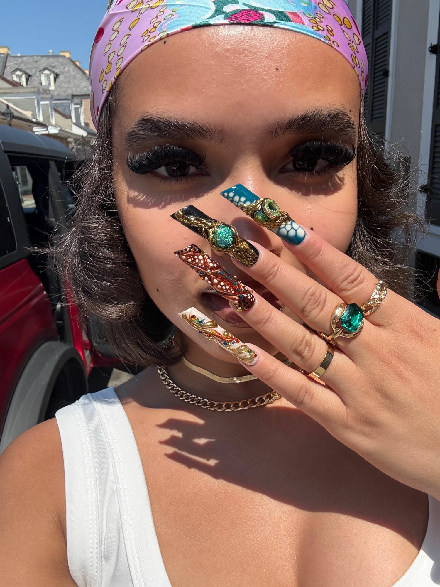 Close-up of a woman with colorful nails, multiple rings, and a headscarf, resting her face on her hand outdoors.