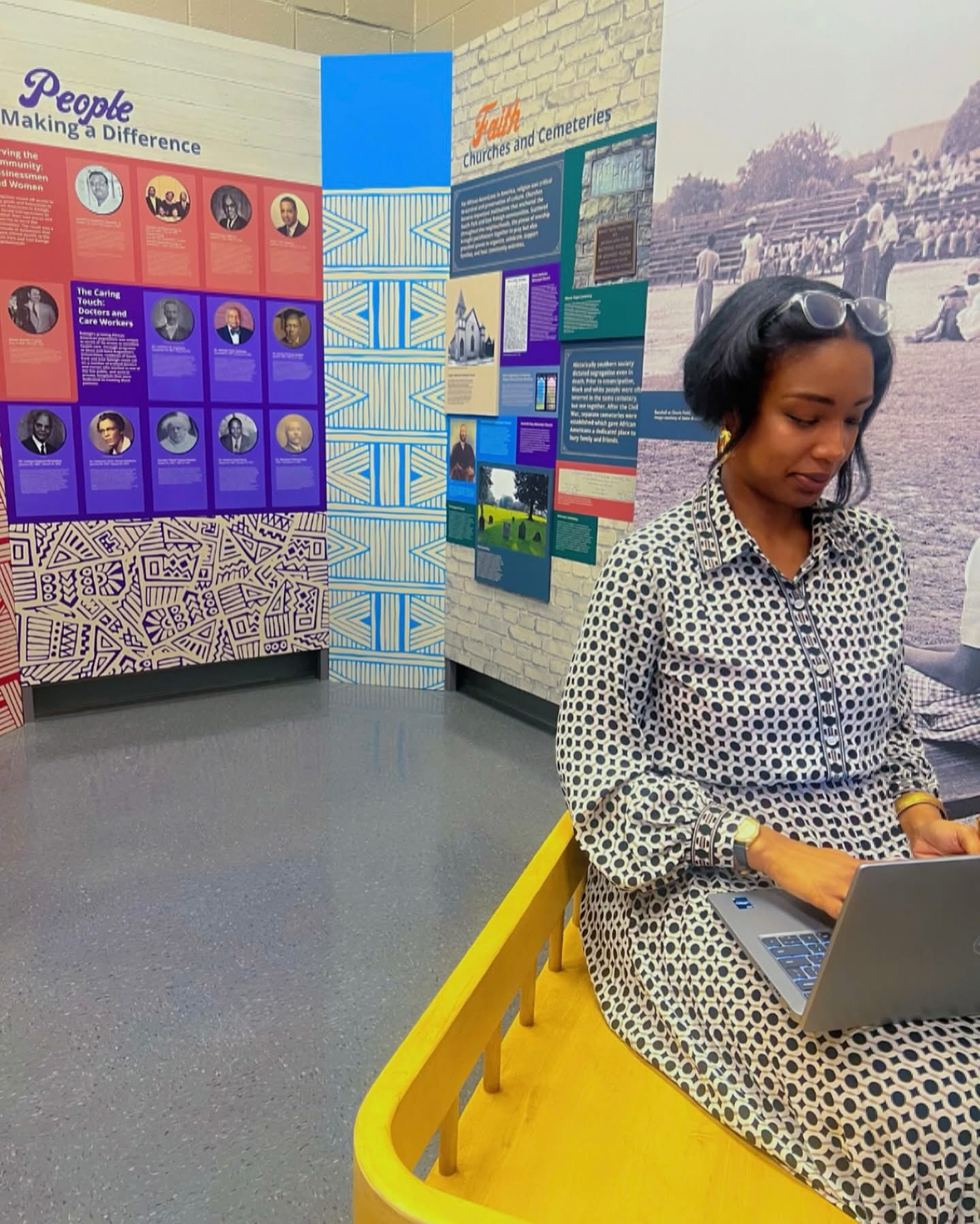 Woman sitting on a yellow bench using a laptop, with informational display boards in the background.