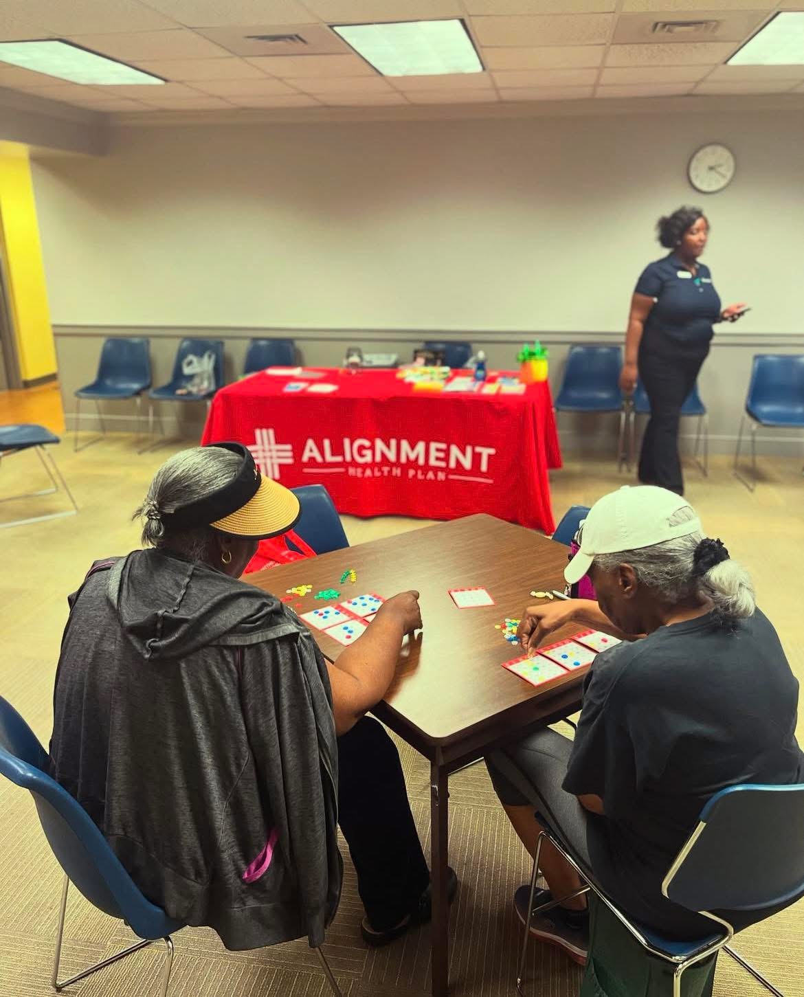 Two women sit at a table playing a card game in a room with chairs, a red tablecloth, and a woman standing in the background.