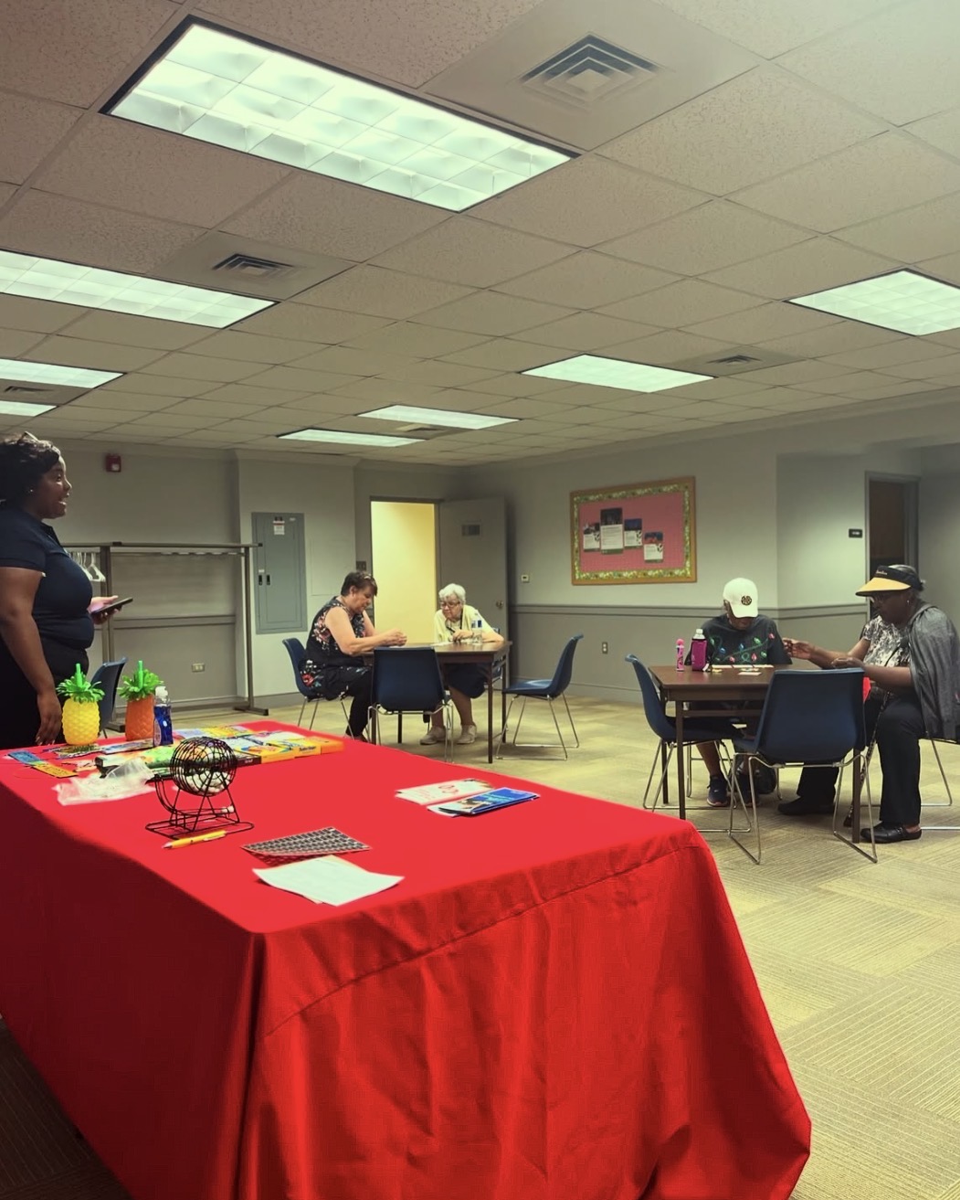 People sitting at tables in a room with a red tablecloth in the foreground, fluorescent ceiling lights, and a colorful wall art.