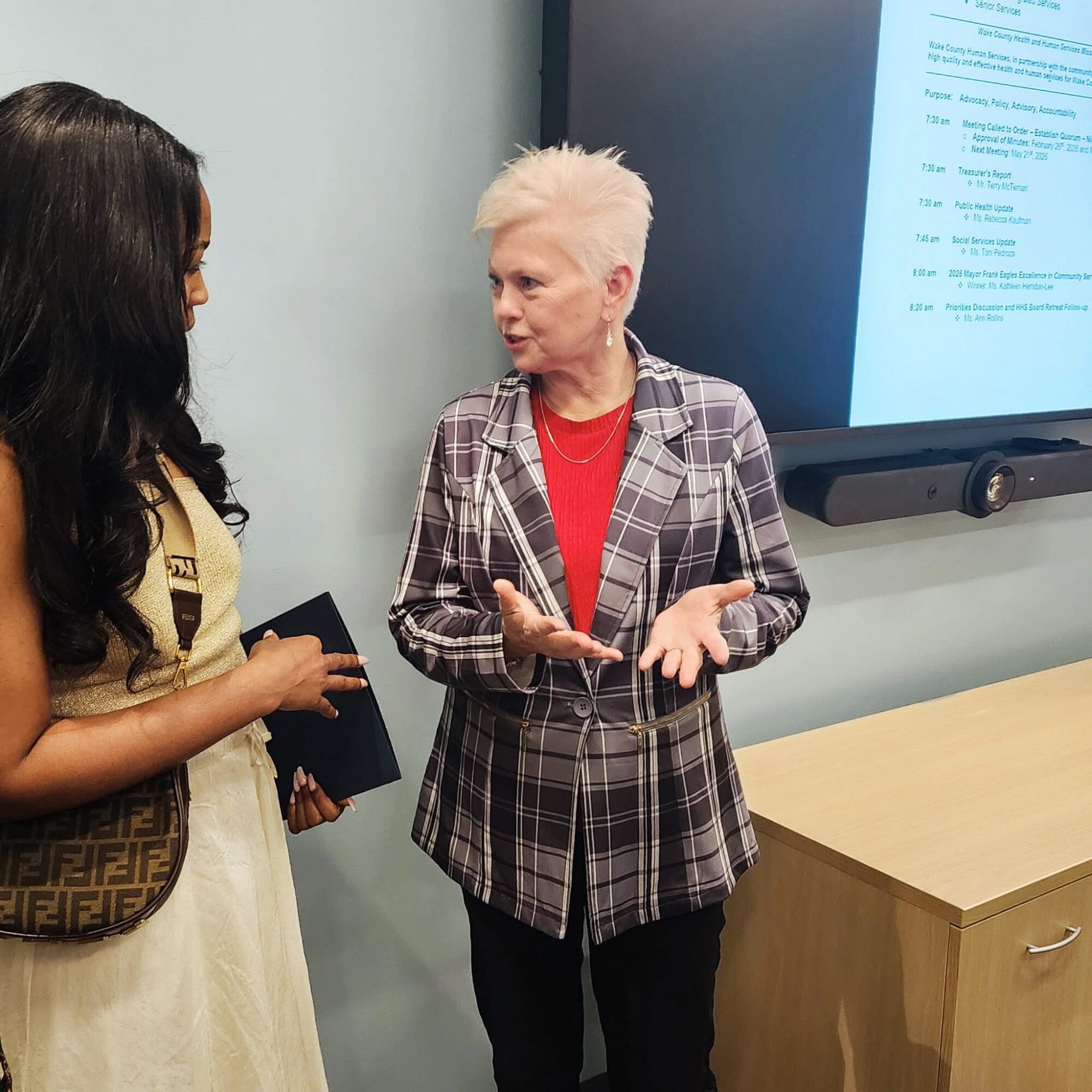 Two women engaged in conversation in a room with a large screen displaying text, one with short white hair and plaid blazer.