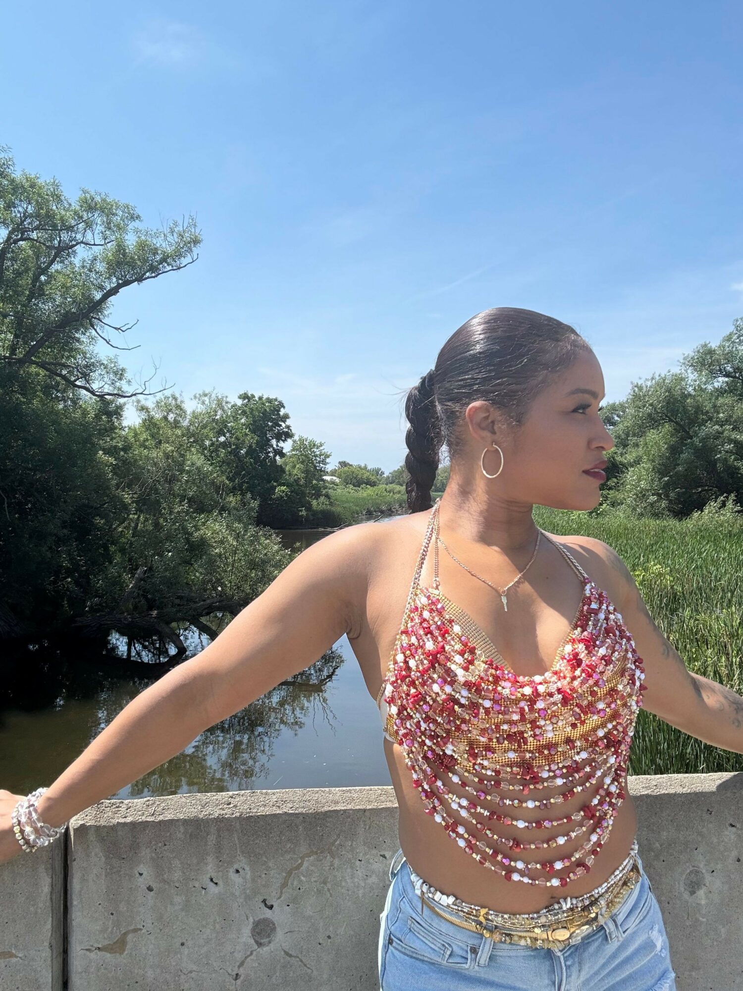 Young woman with braided hair wearing a beaded top and denim shorts, standing outdoors with trees and sky in background.