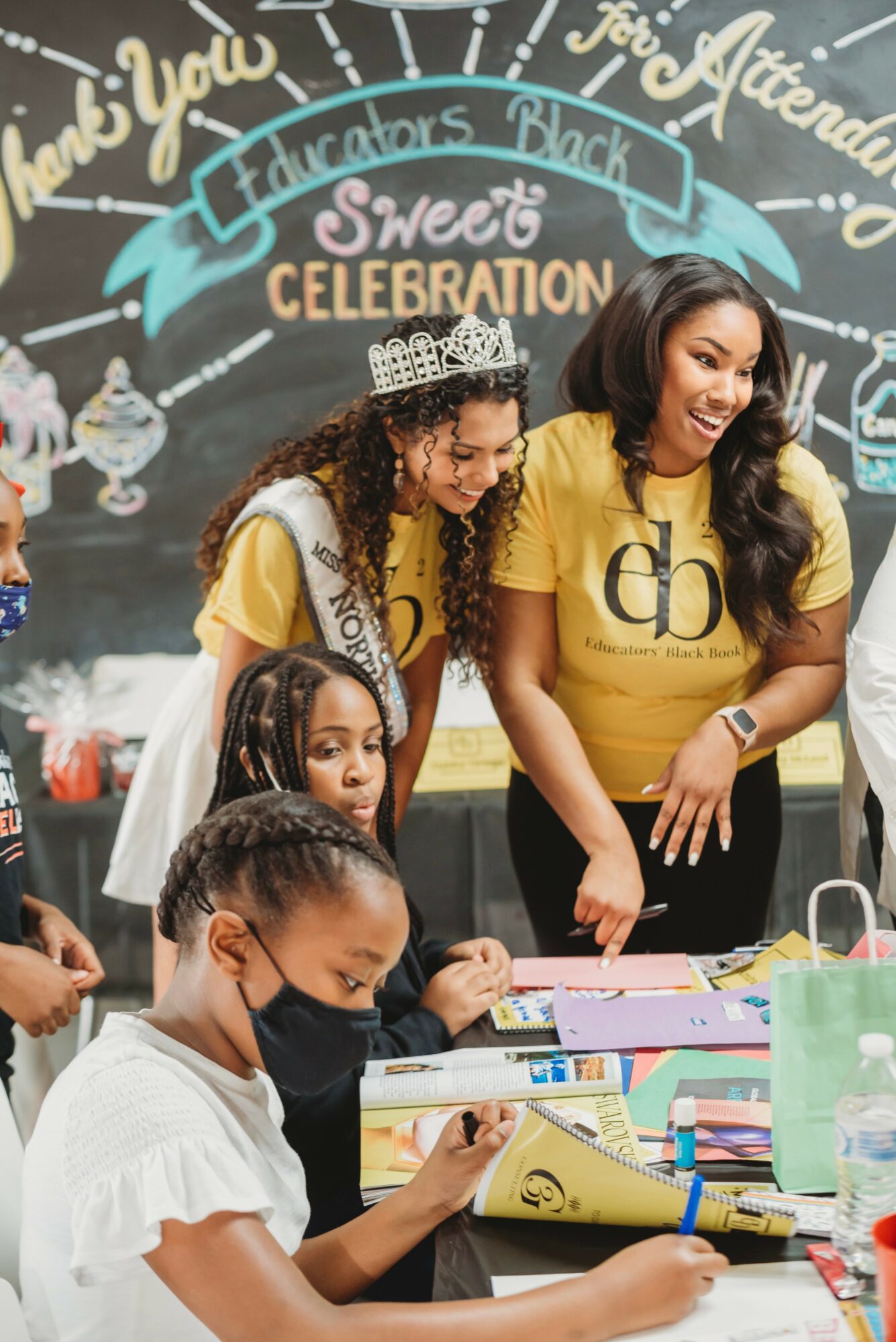 Two women and children at a celebration table with colorful decorations and a chalkboard background, smiling and engaging in activities.