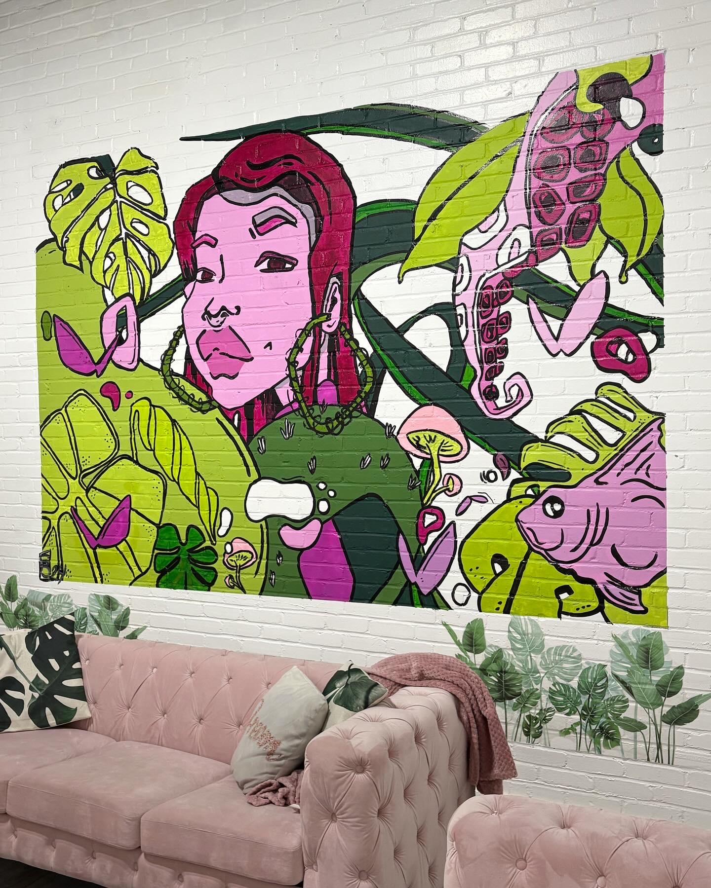 Colorful mural of a woman surrounded by plants and animals, with pink, green, and purple tones, above a pink sofa.