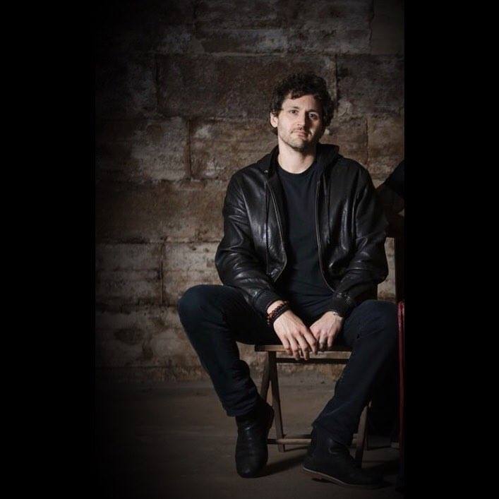 Young man with dark curly hair and beard, wearing a black leather jacket, sitting on a chair against a textured wall.