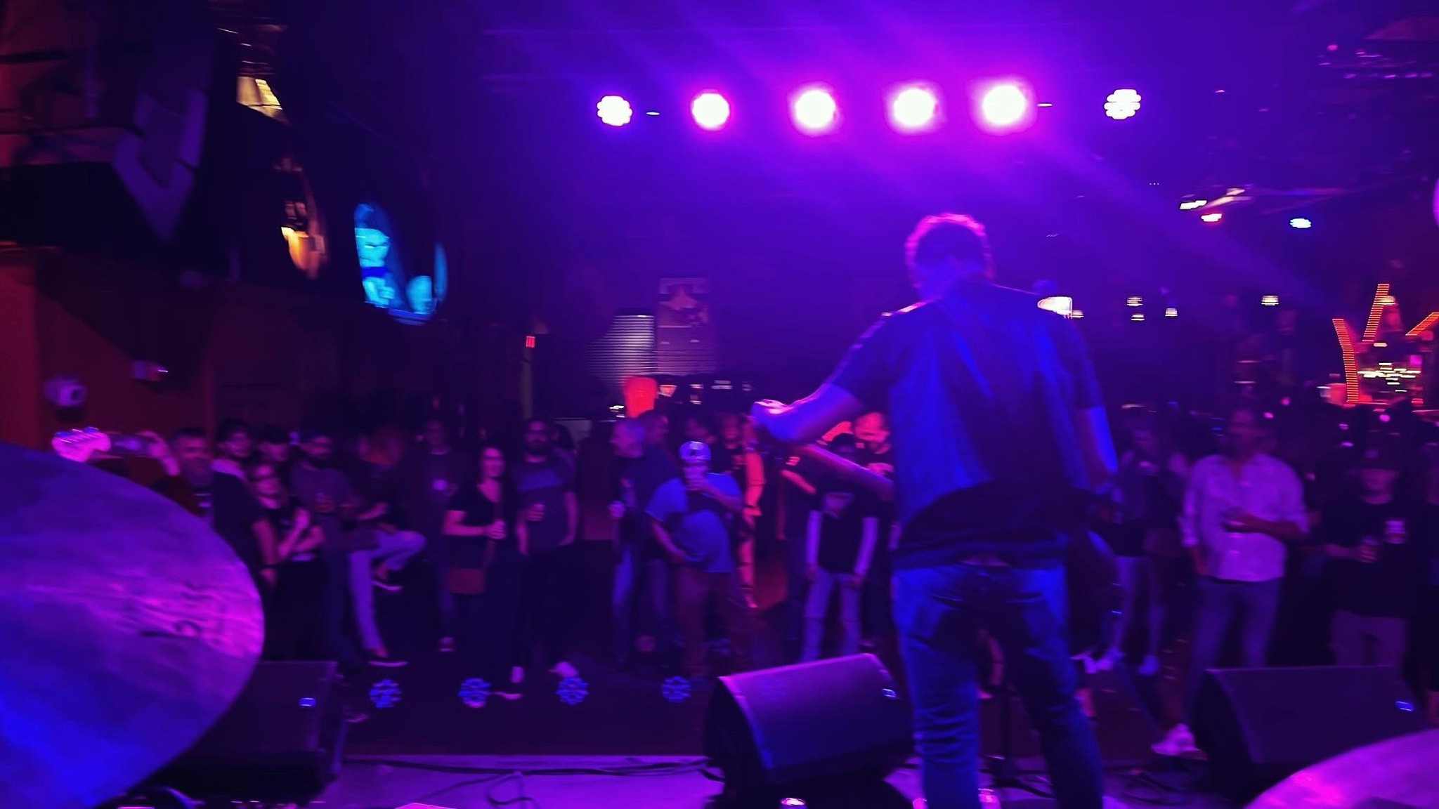 Performer on stage with a microphone, facing a crowd in a dimly lit venue with purple lighting.