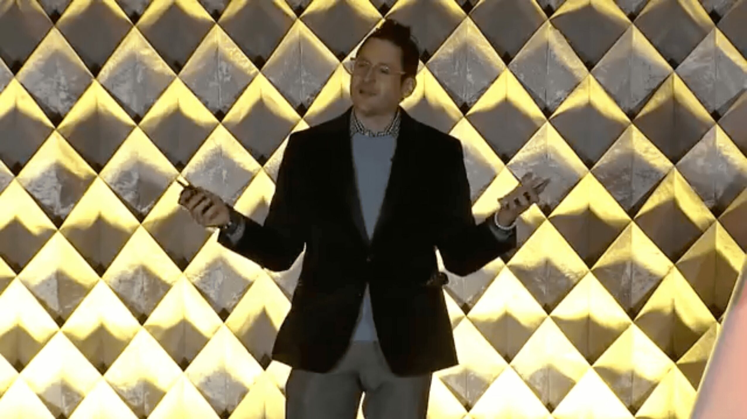 Person in a suit standing in front of a geometric patterned wall, holding a phone in each hand, smiling.