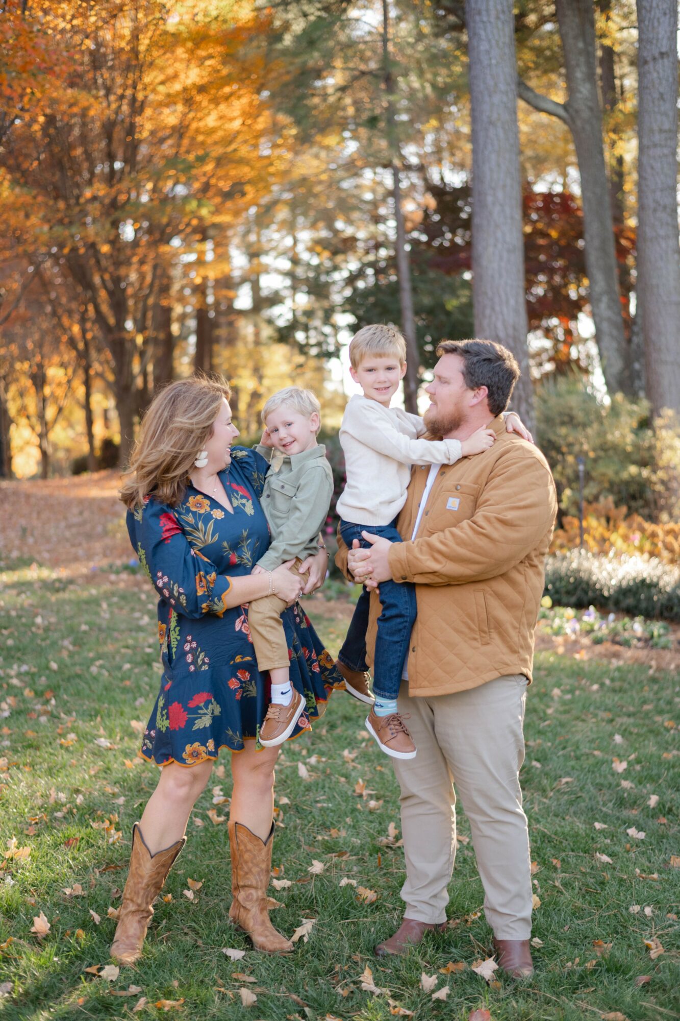 Family of four outdoors in autumn, with trees and fallen leaves, smiling and playing together.
