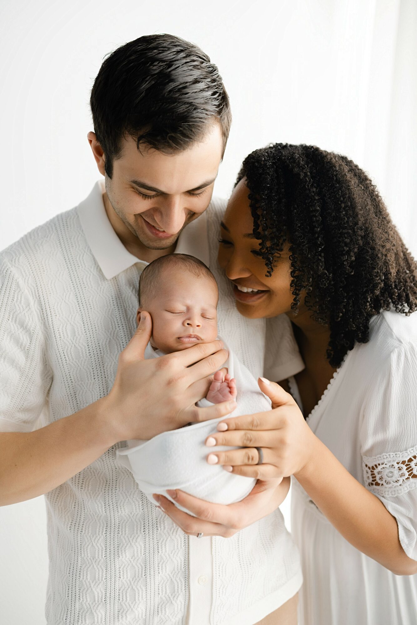 Young couple holding a sleeping baby, smiling and looking at the baby, in a bright room with white curtains.