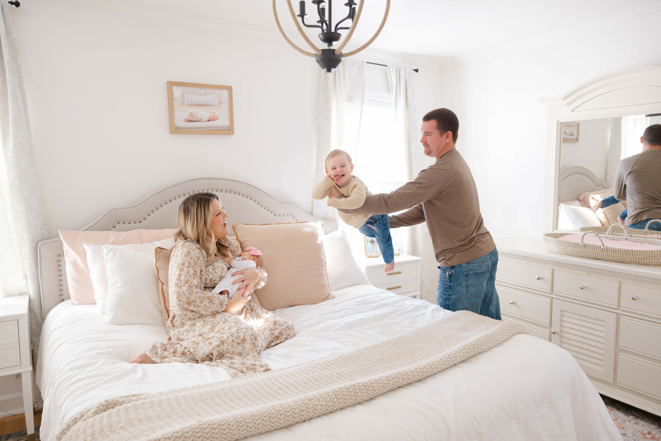 A man hands a baby to a woman sitting on a bed in a bright bedroom with white furniture and curtains.