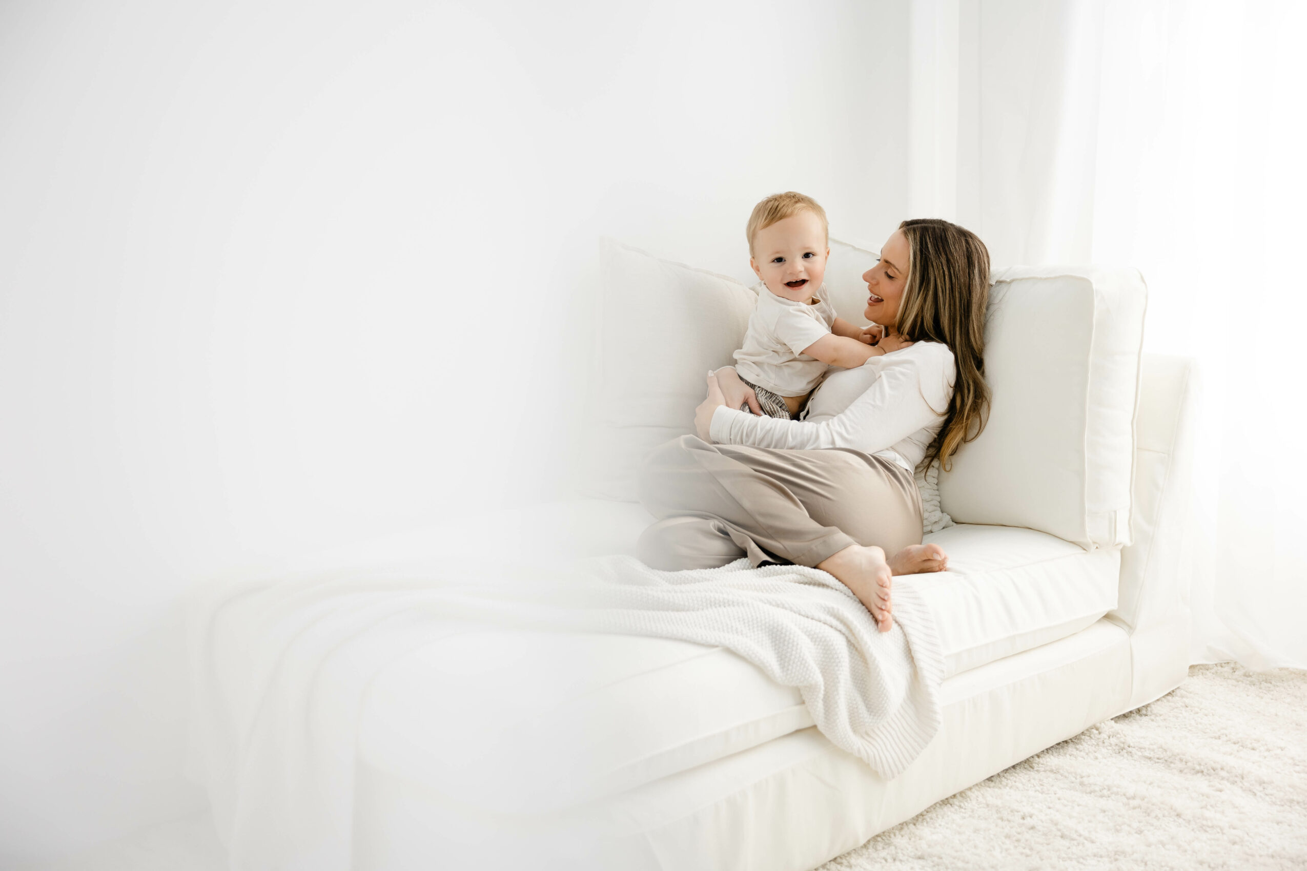 Woman and baby sitting on a white sofa in a bright room, woman holding the baby, both smiling.