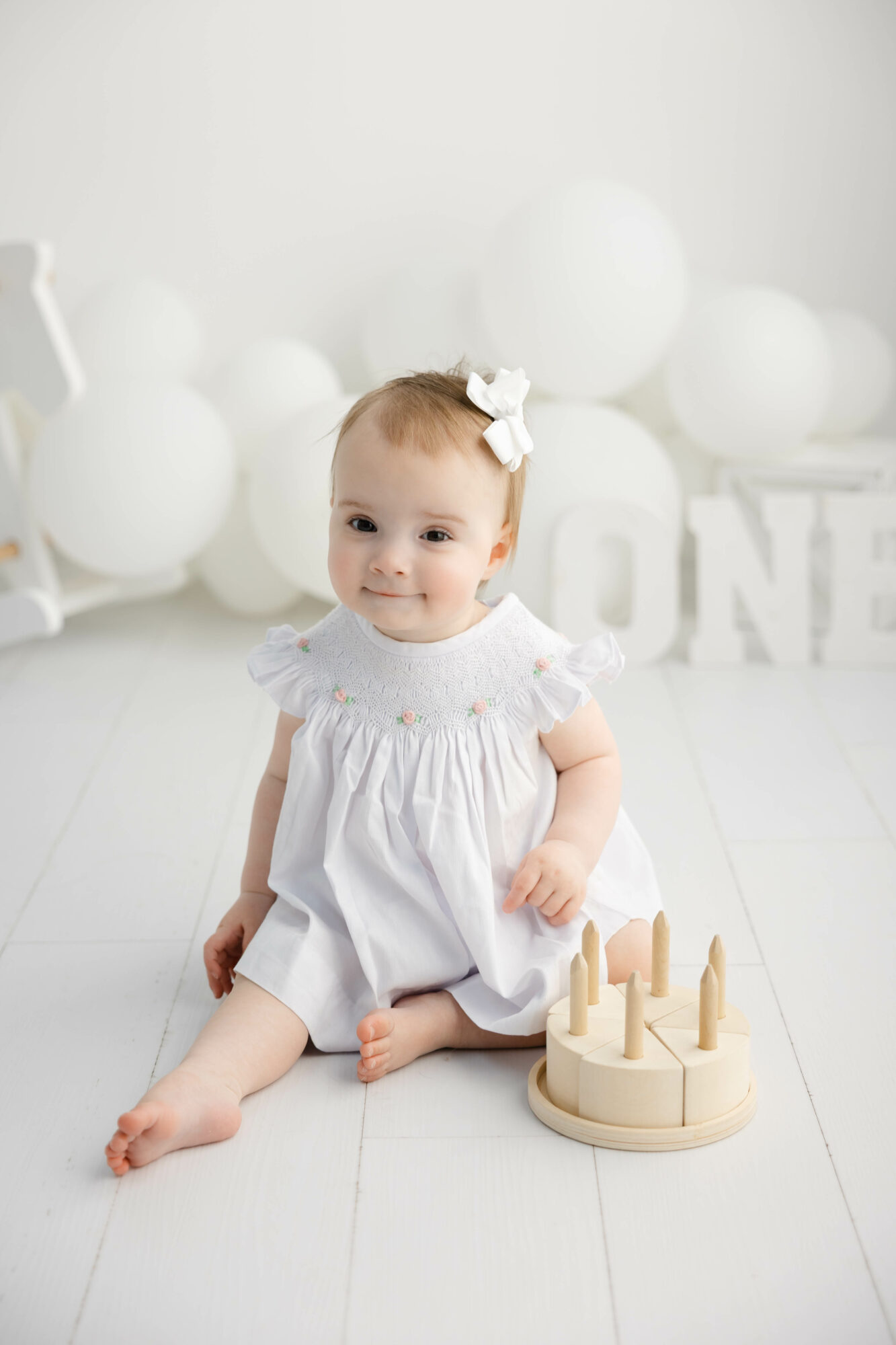 Baby girl sitting on white floor with wooden castle toy, wearing white dress and bow, in a bright room with balloons and decorations