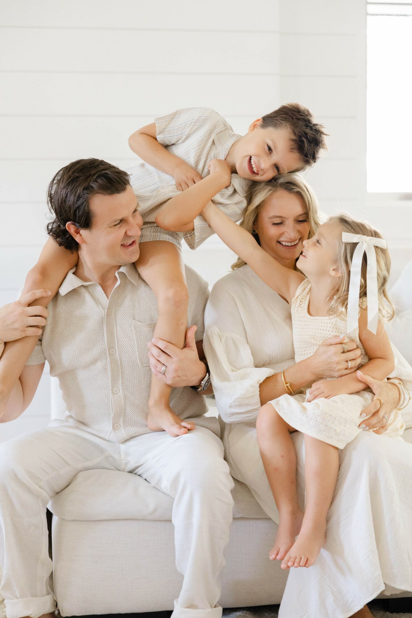 Family of four playing and laughing together on a white sofa in a bright room, with a window in the background.