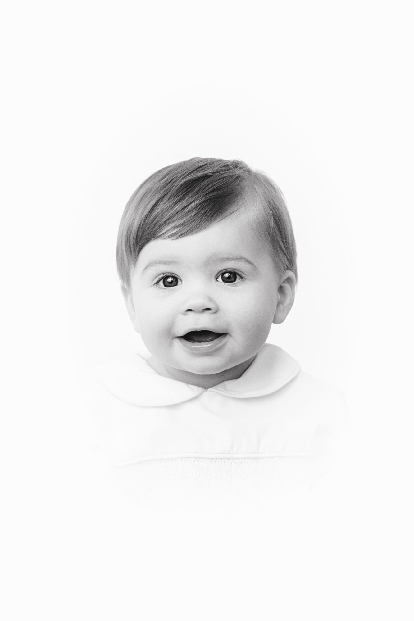 Baby with short hair, smiling, wearing a collared shirt, in black and white, centered, plain background.