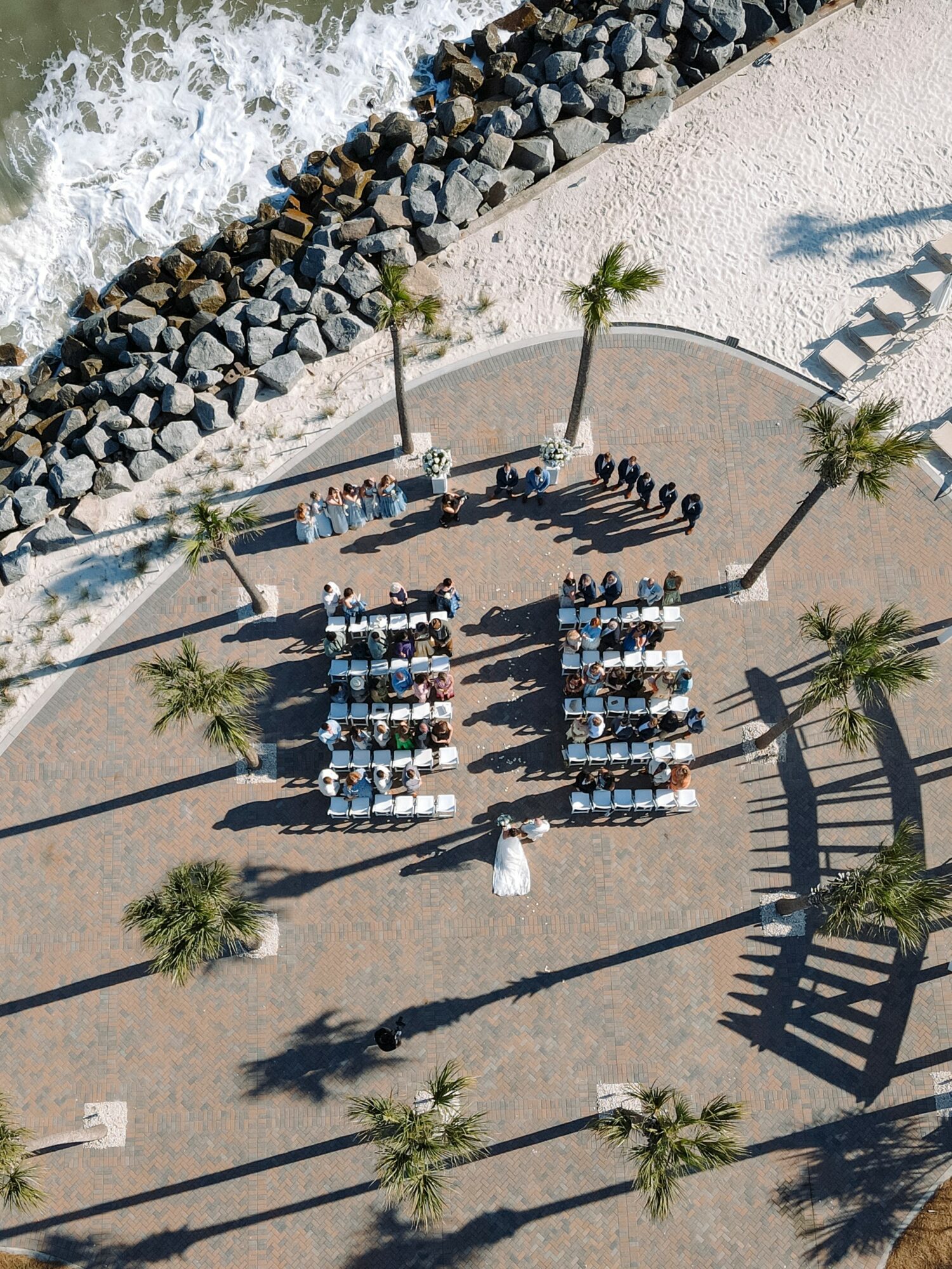 People sitting at tables on a beachside promenade with palm trees, near rocks and water, viewed from above.