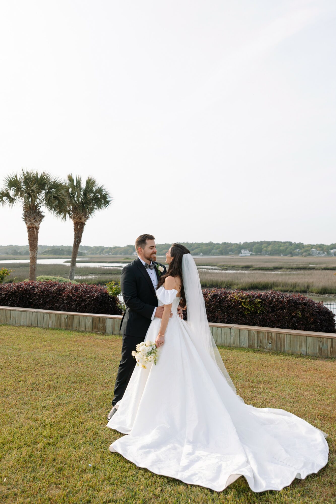 A bride and groom stand outdoors on grass, holding hands and looking at each other, with palm trees and a body of water in the background.