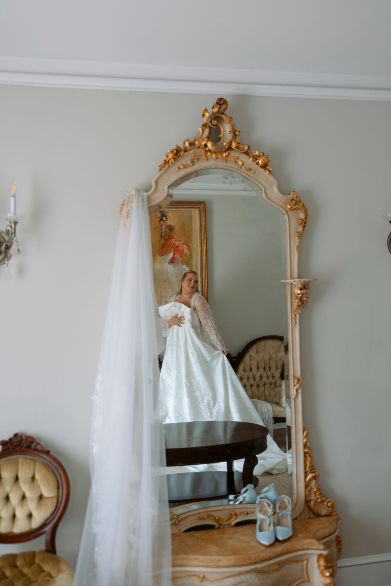 Person in a white dress taking a photo in a large ornate mirror with gold accents, reflected in a room with a painting and furniture.