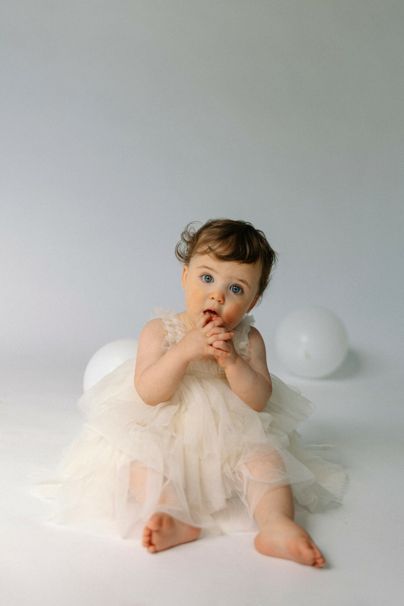 Baby girl sitting on a white surface with two white balloons behind her, wearing a light-colored dress, holding her hands near her mouth.