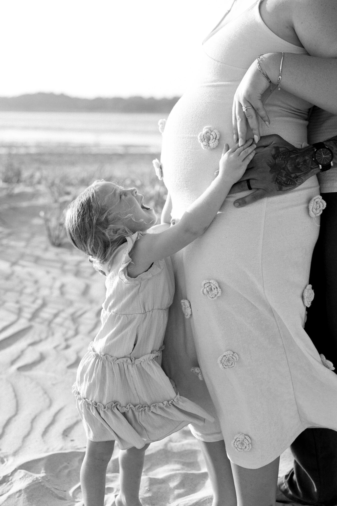 Child touching pregnant woman's belly outdoors, with a beach and water in the background, in black and white.