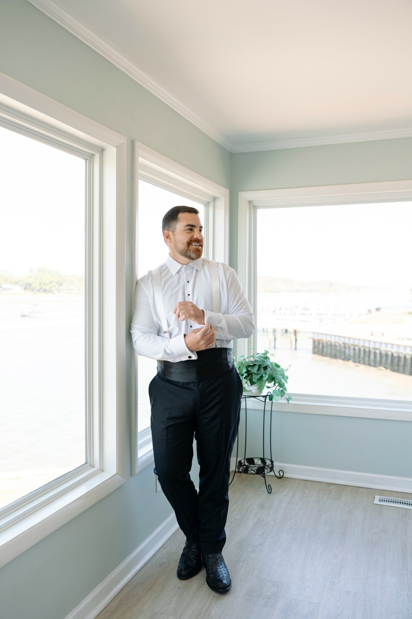 Man in white shirt and dark pants standing by windows in a bright room with light green walls and a potted plant.