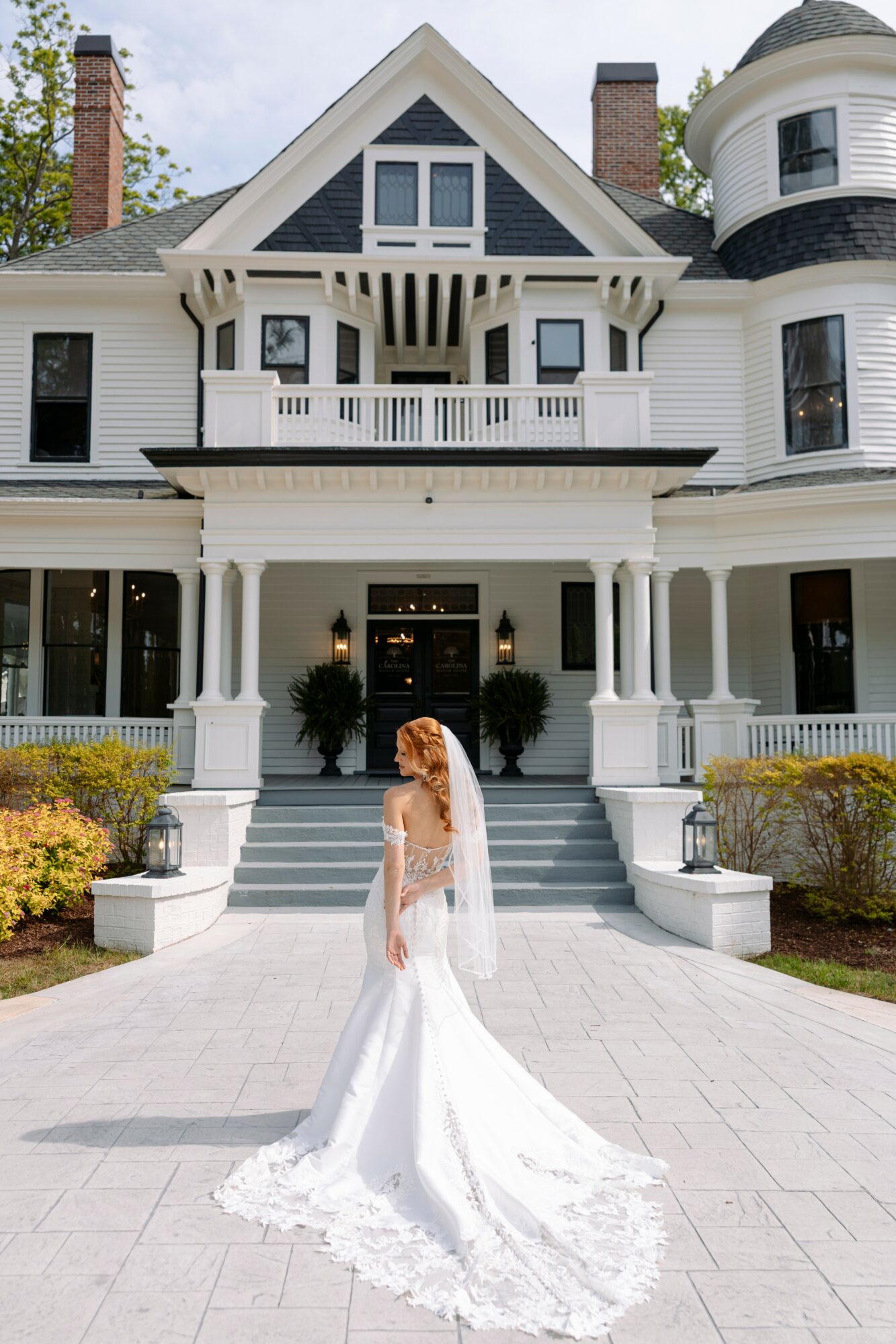 Woman in wedding dress standing on steps in front of a large, historic house with a porch and turret.