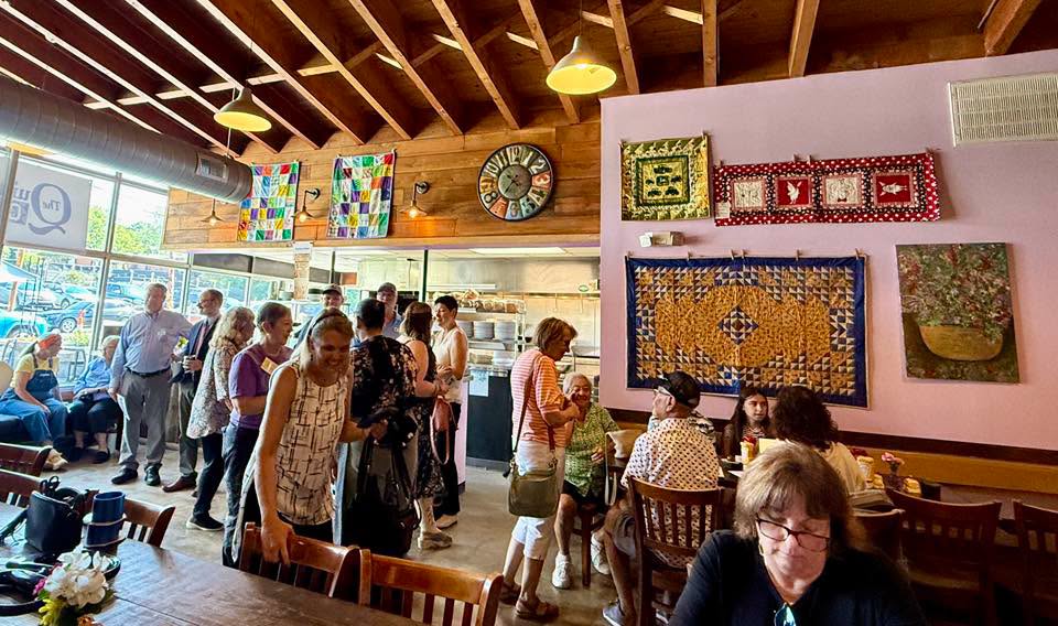 People gather inside an art gallery with colorful textile artworks on pink walls and wooden ceiling beams.