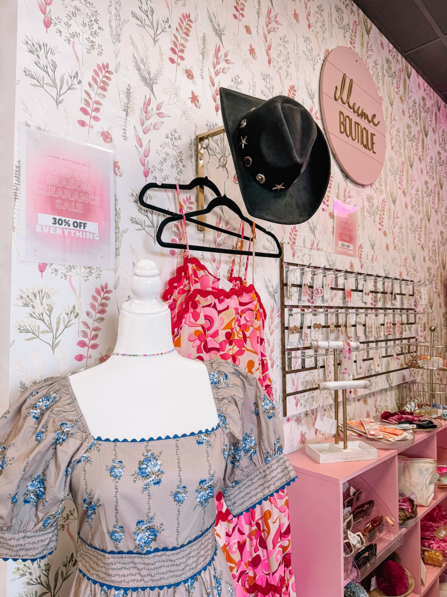 Display of hats, clothing, and accessories on a wall with floral wallpaper, including a mannequin with a dress, and pink shelves.