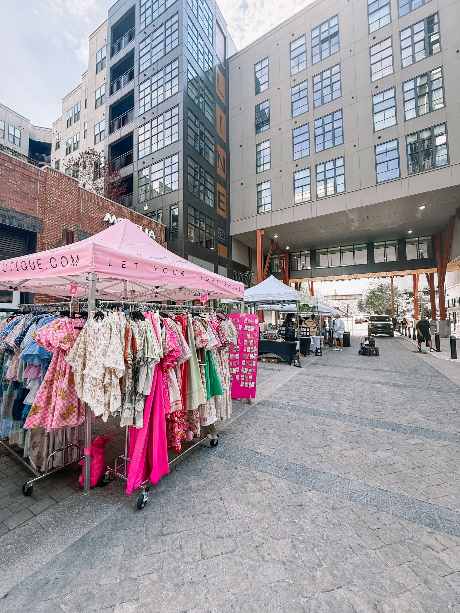 Outdoor market with clothing stalls and tents in an urban area with tall buildings, paved walkway, and people in the background.