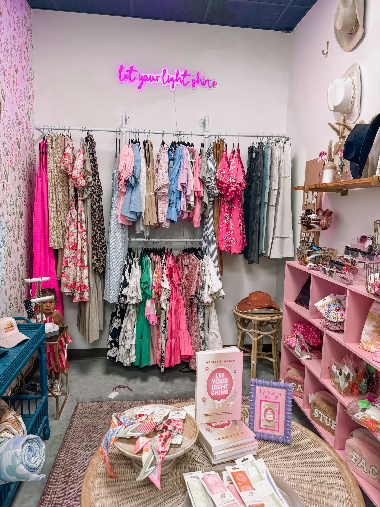 Clothing rack with colorful dresses and tops, pink shelves with accessories, neon sign, and a table with products inside a boutique.