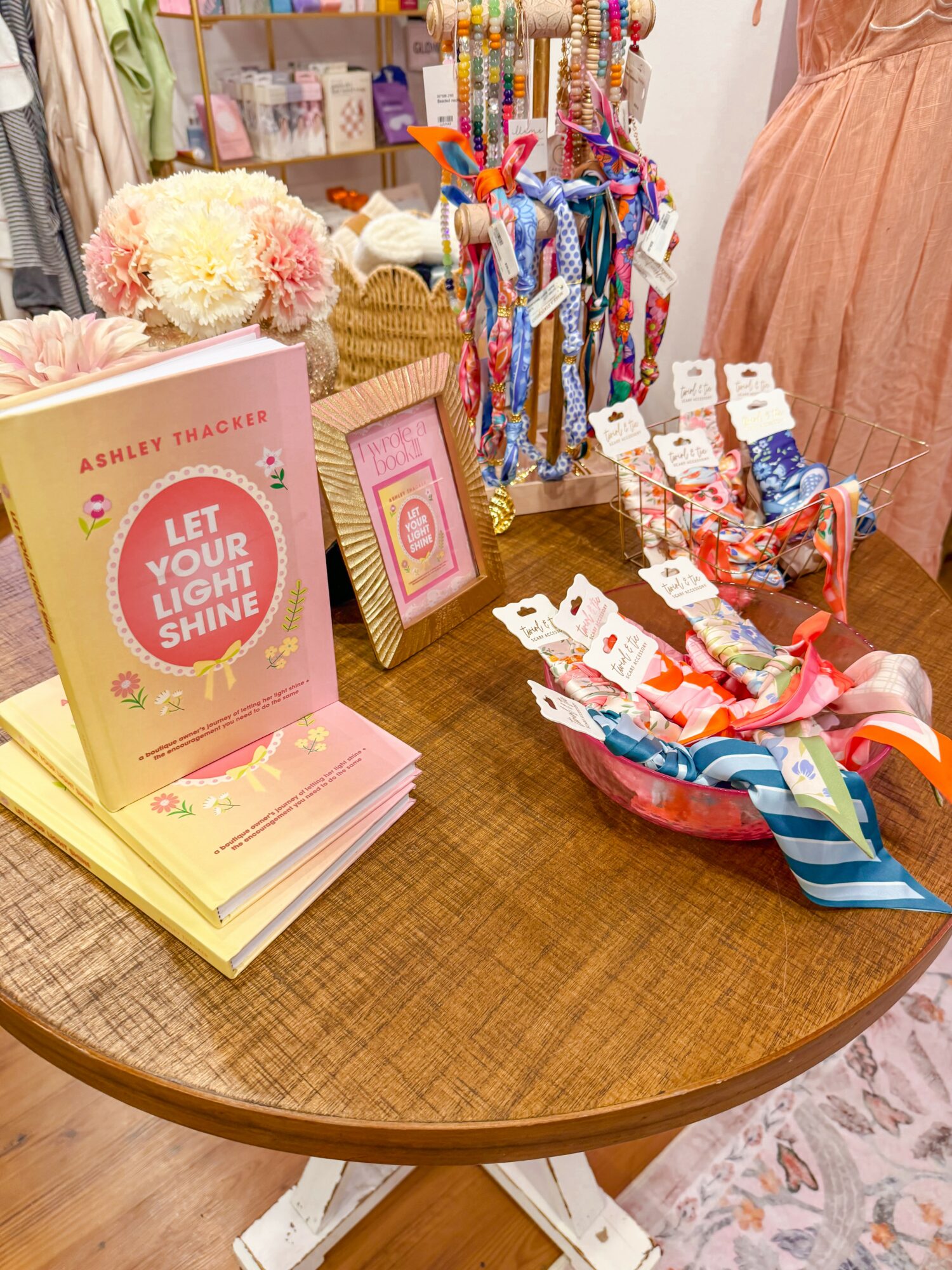 Display table with books, jewelry, and accessories in a store, with a person in a peach dress nearby.
