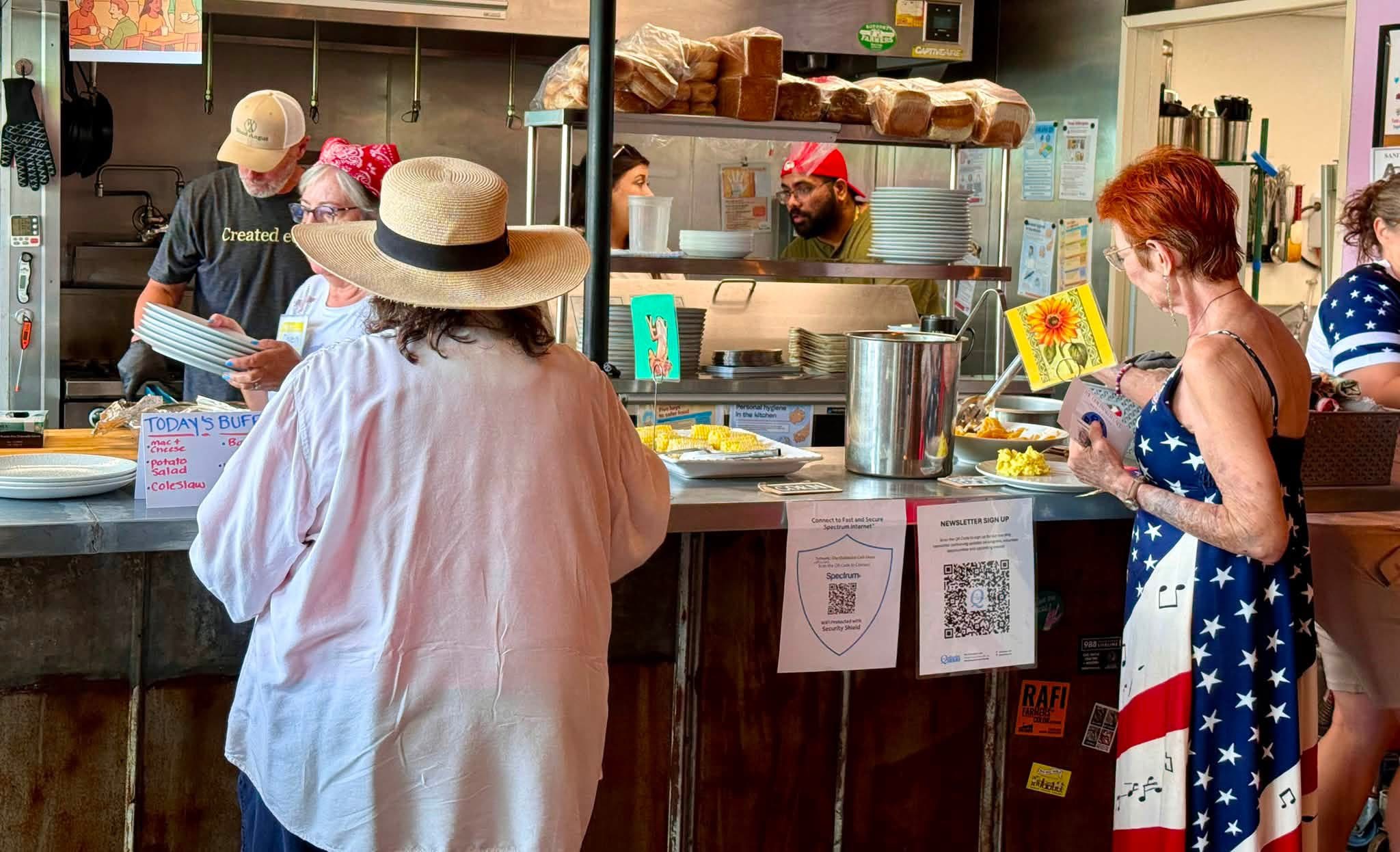 People ordering food at a counter in a casual eatery, with staff preparing food behind the counter, and signs hanging above.