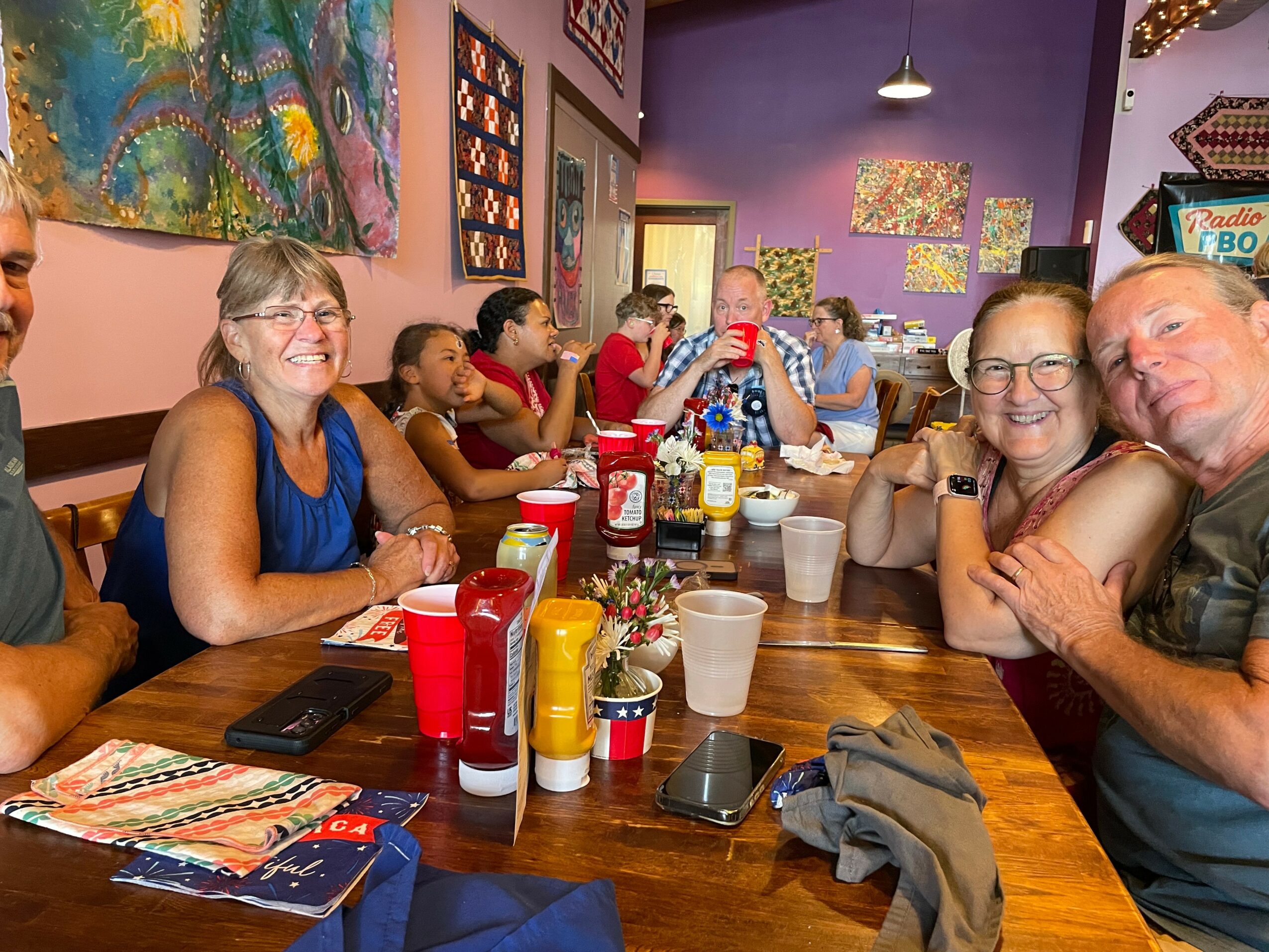 Group of people sitting at a restaurant table, smiling and enjoying a meal, with colorful artwork on the walls.