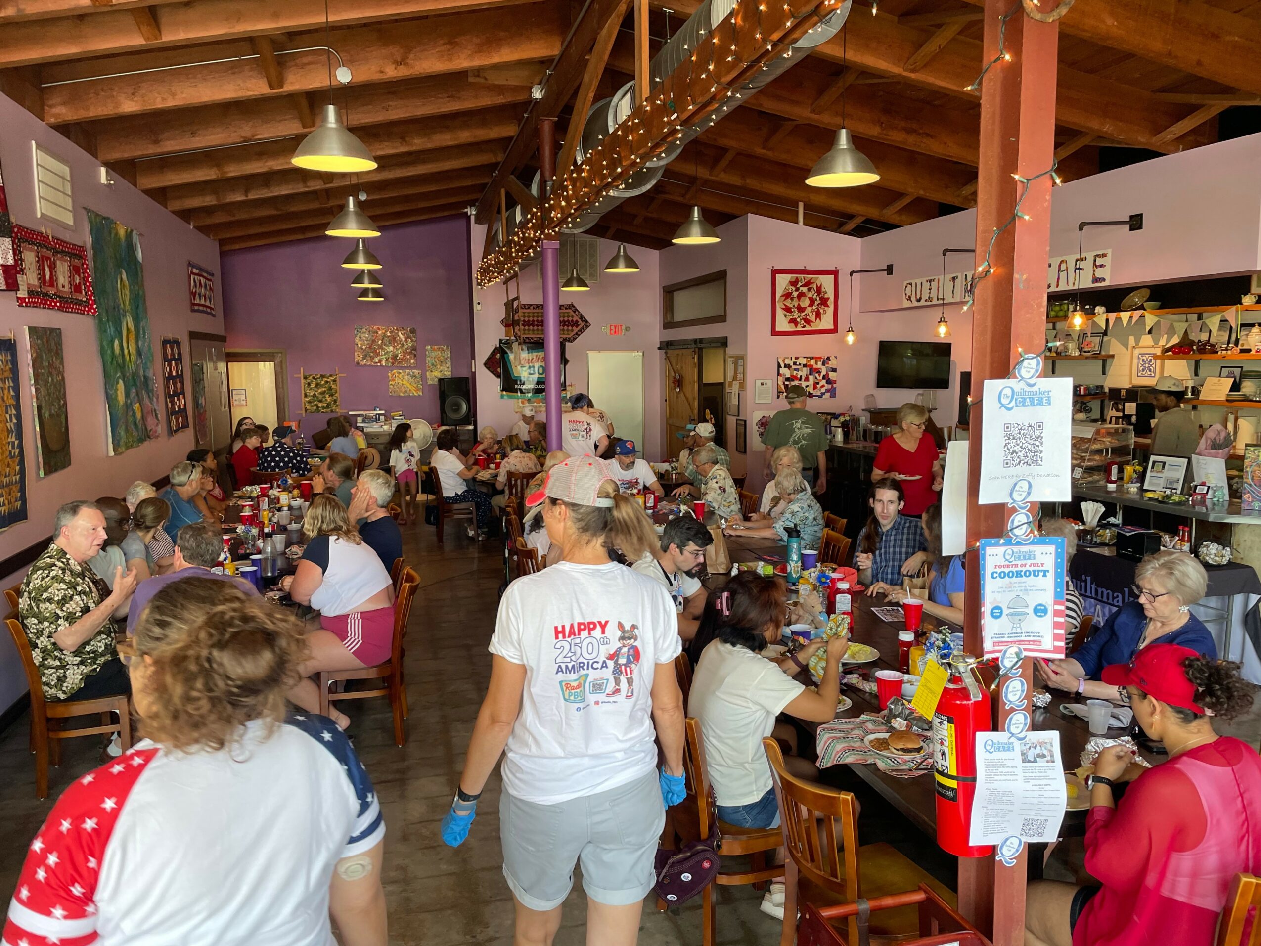 Crowded indoor space with people sitting at tables, colorful walls, and wooden ceiling beams.