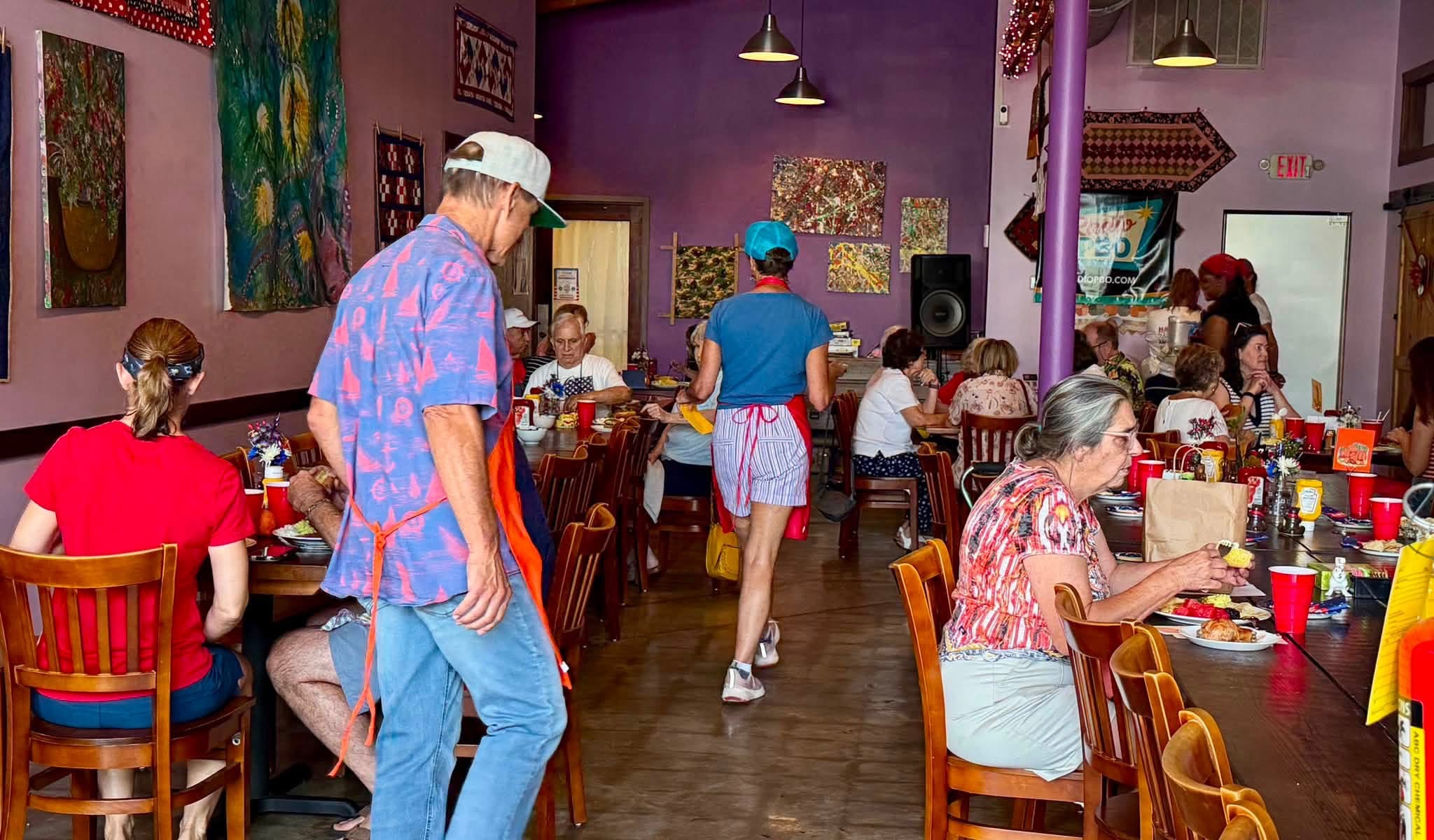 People dining in a colorful restaurant with purple walls, wooden chairs, and hanging lights, some wearing casual summer clothing.