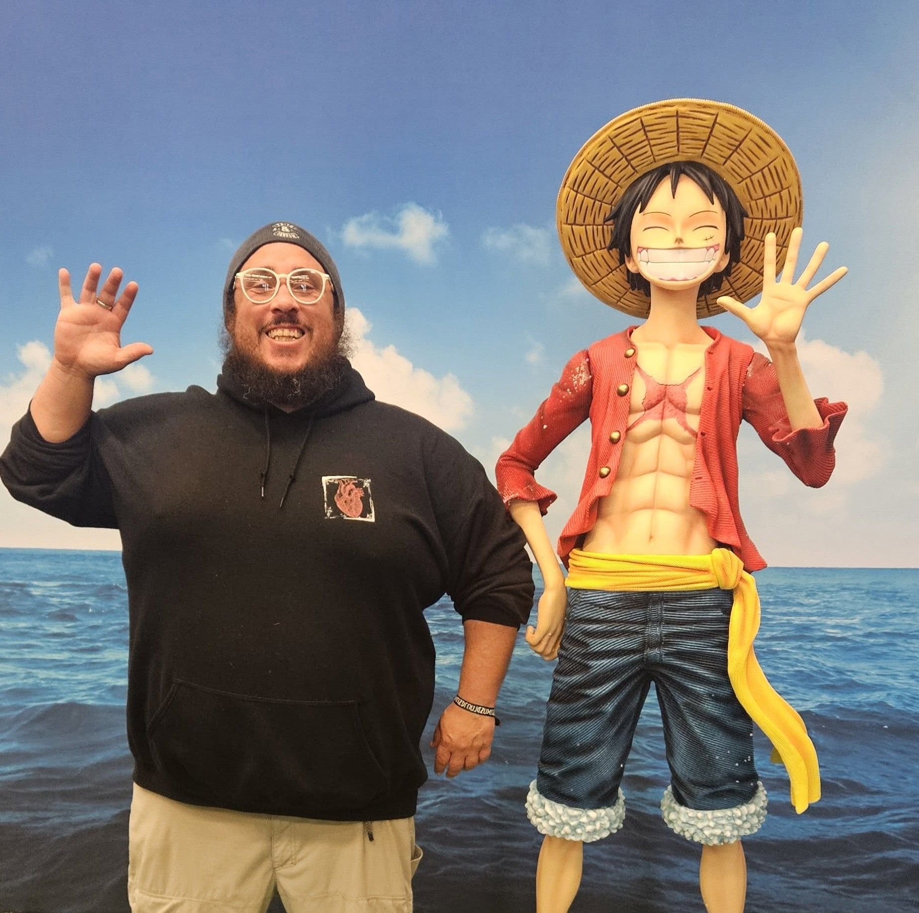 Person with glasses and beard smiling and waving next to a large animated character on a beach, ocean in background.