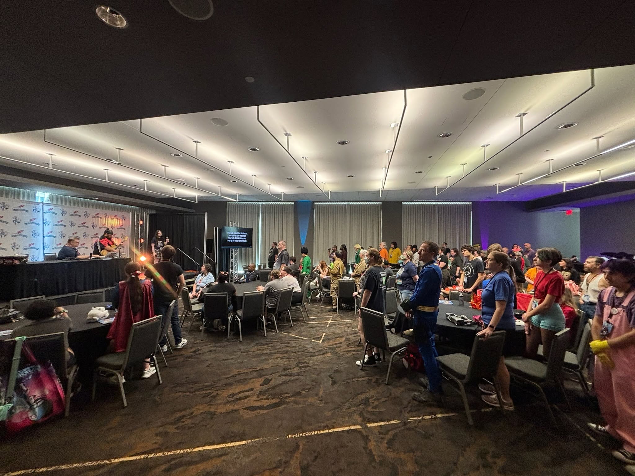 People gathered in a large room with tables and chairs, some standing and some sitting, with a stage and curtains in the background.